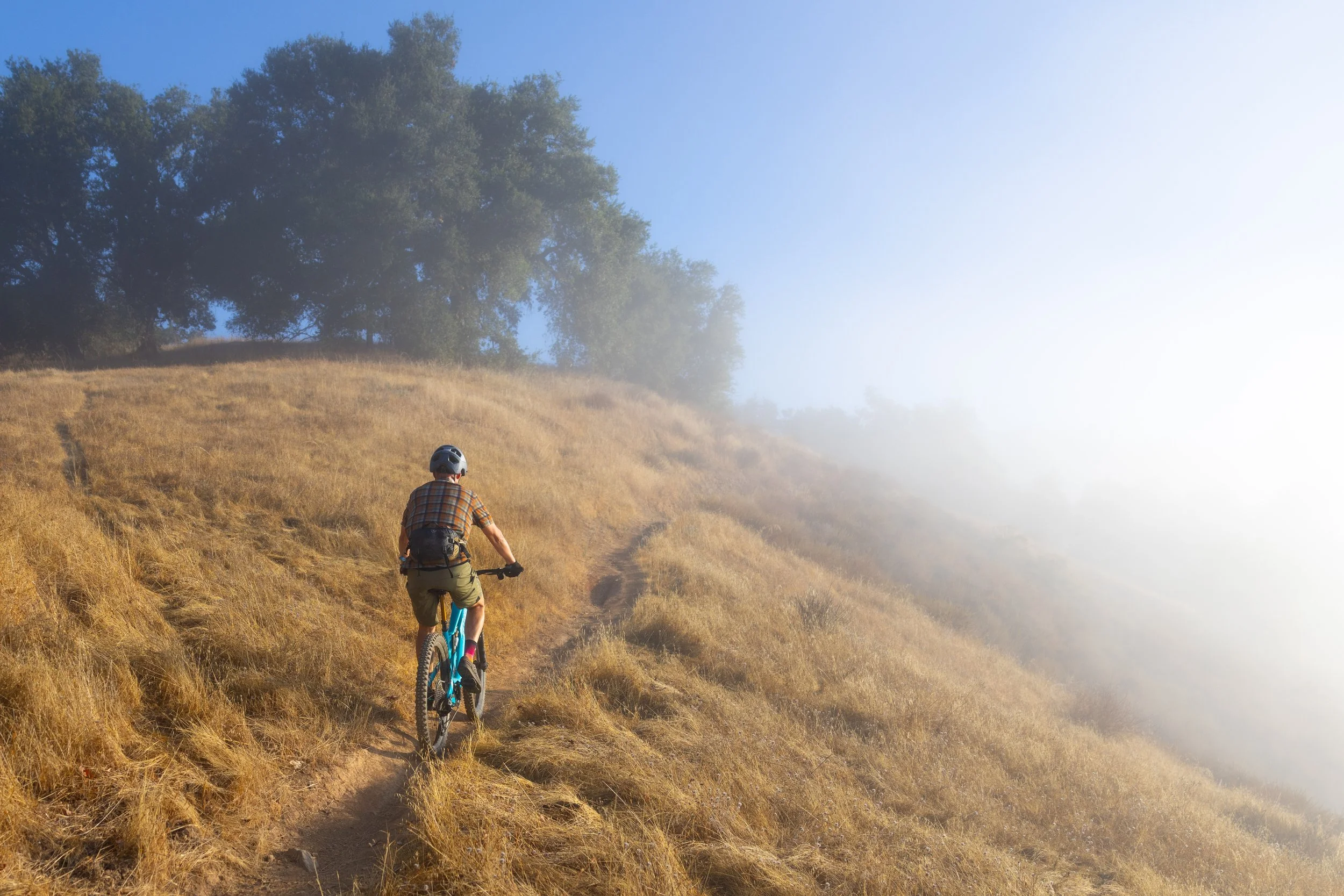 A person riding a mountain bike on a narrow dirt trail on a dry, grassy hillside. The rider is wearing a helmet and a plaid shirt, and there are trees at the top of the hill with fog or mist in the background.