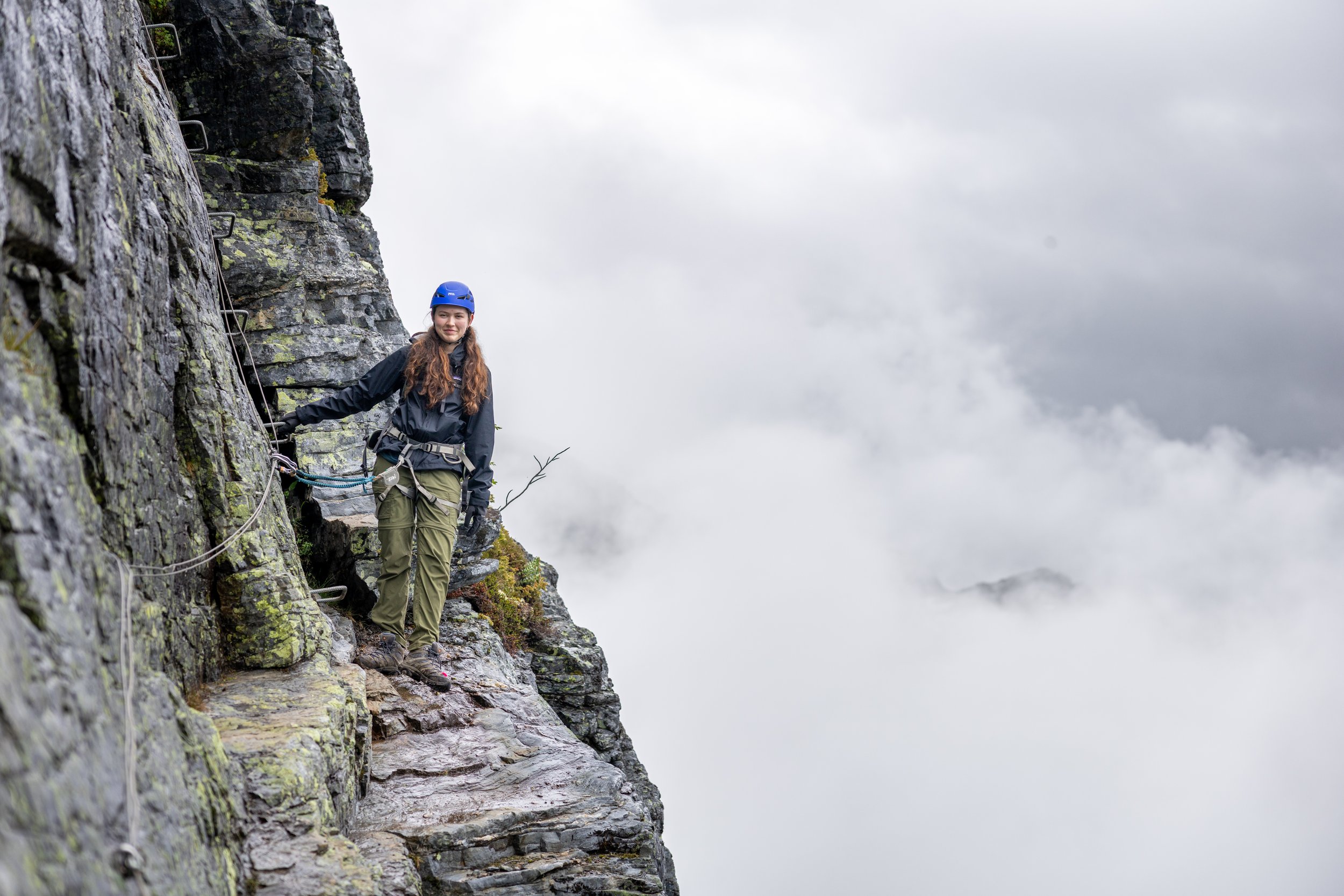 A woman wearing a blue helmet and outdoor gear climbing a rocky mountain trail with safety ropes, surrounded by clouds.