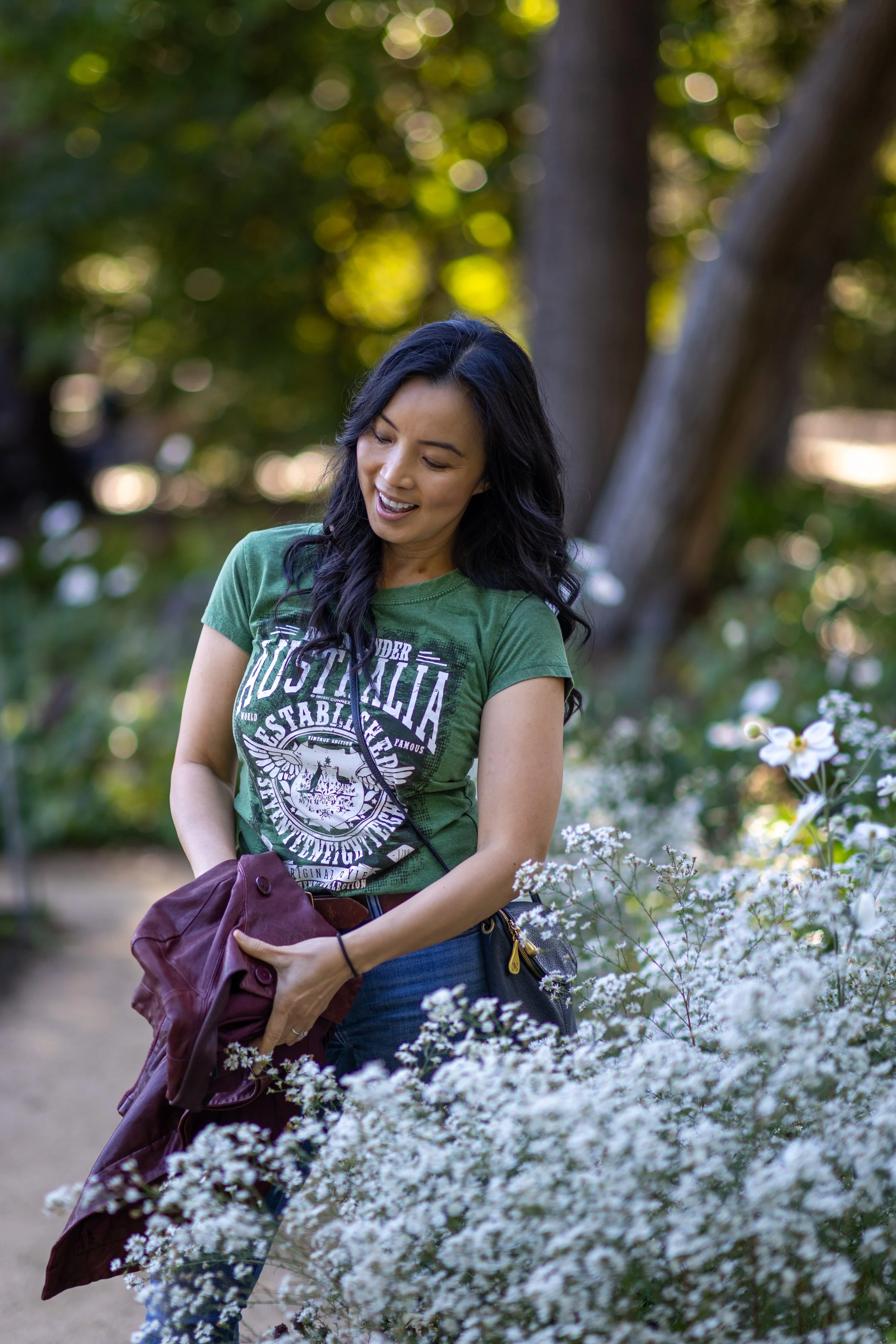A woman with long black hair in a green T-shirt with white graphics, standing outdoors among white flowers and trees, smiling and holding a maroon jacket.