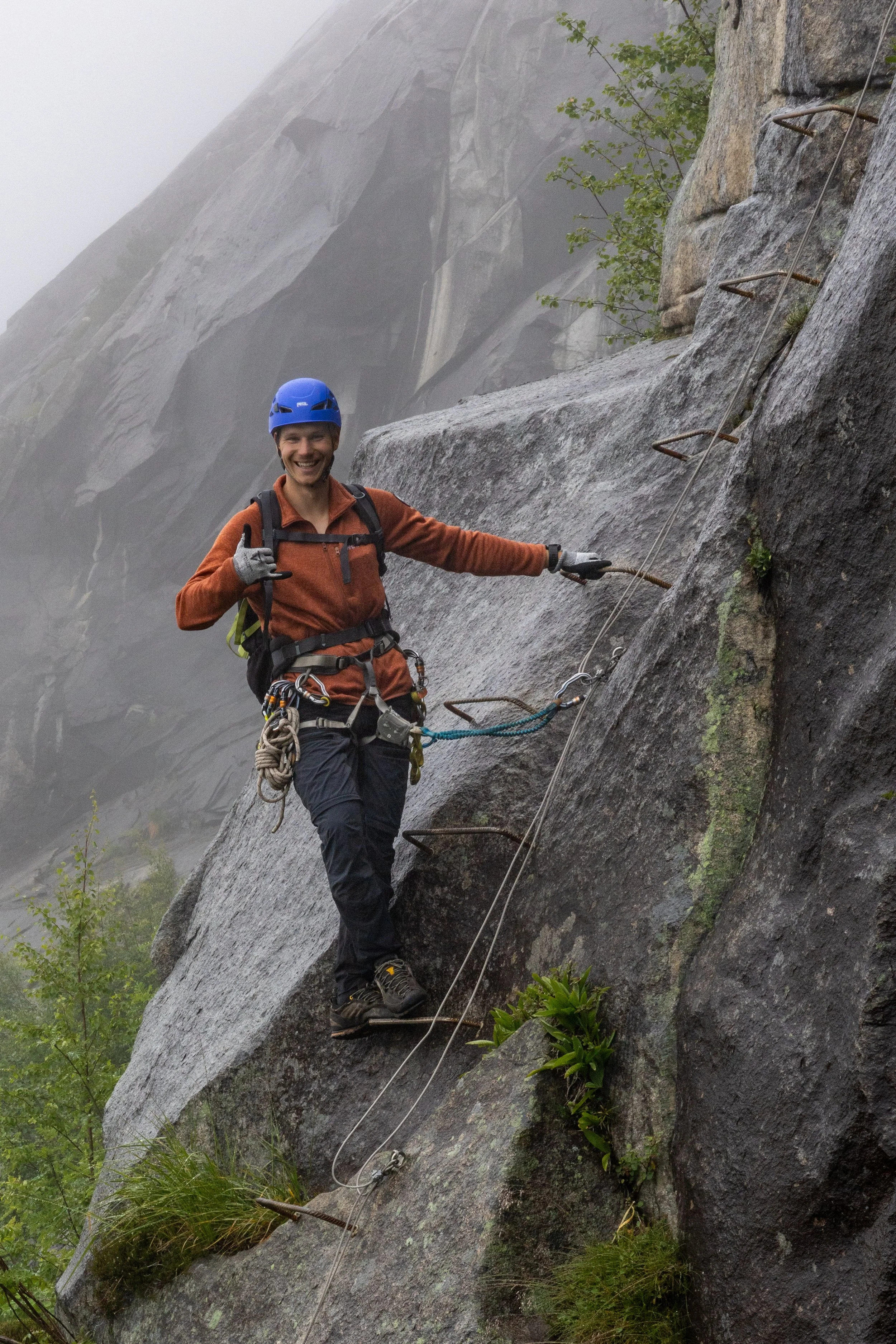 A person wearing a blue helmet, orange jacket, and black pants climbing a steep rock face using metal rungs and safety gear.