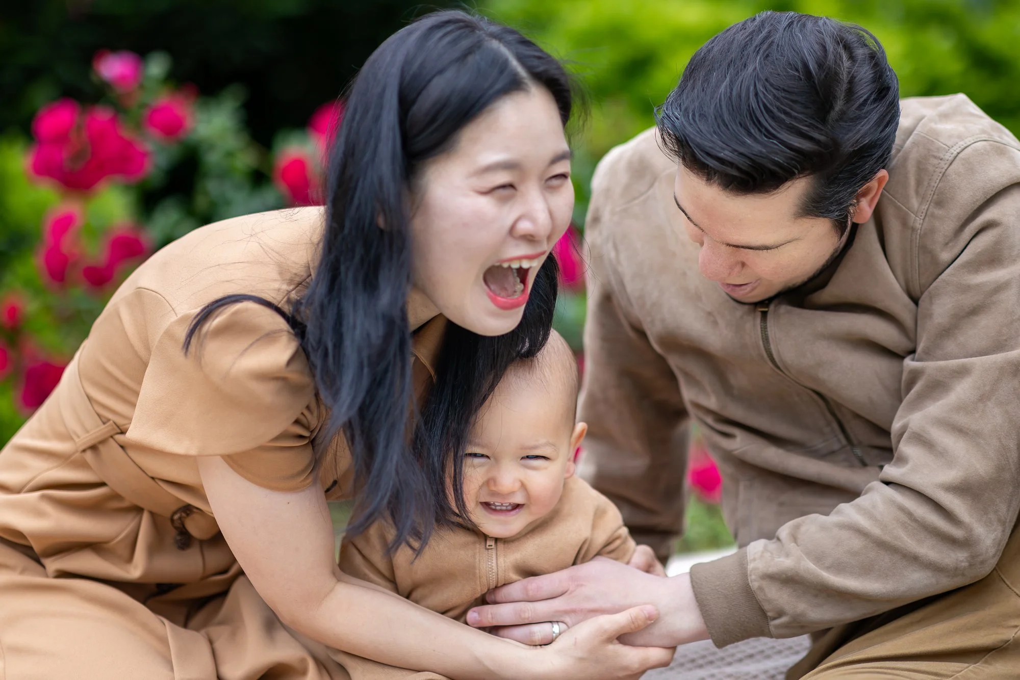 A happy family outside with two adults and a child, all smiling and laughing, surrounded by green grass and pink flowers.