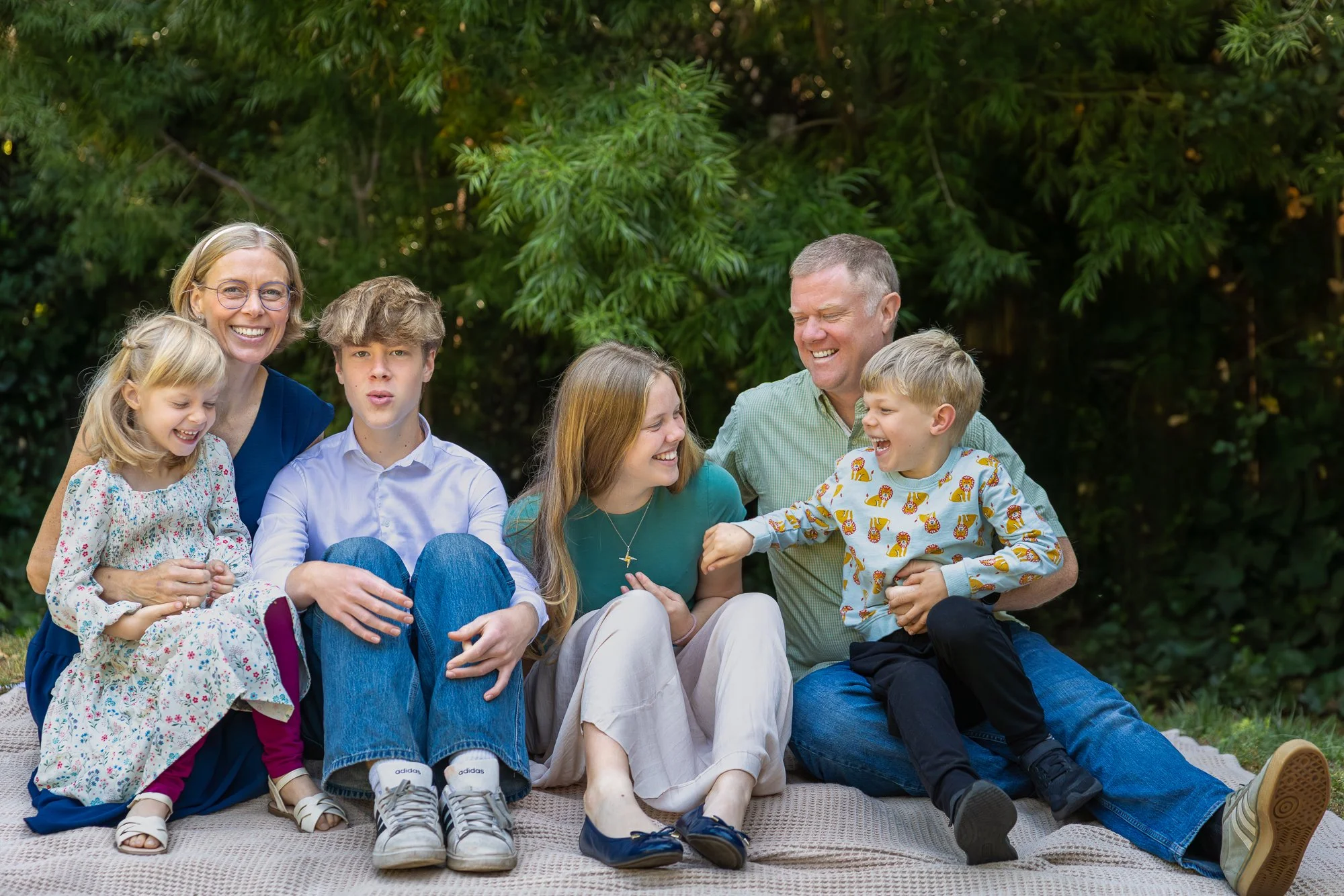 Family of six sitting outdoors on a blanket, laughing and smiling together with a lush green background.