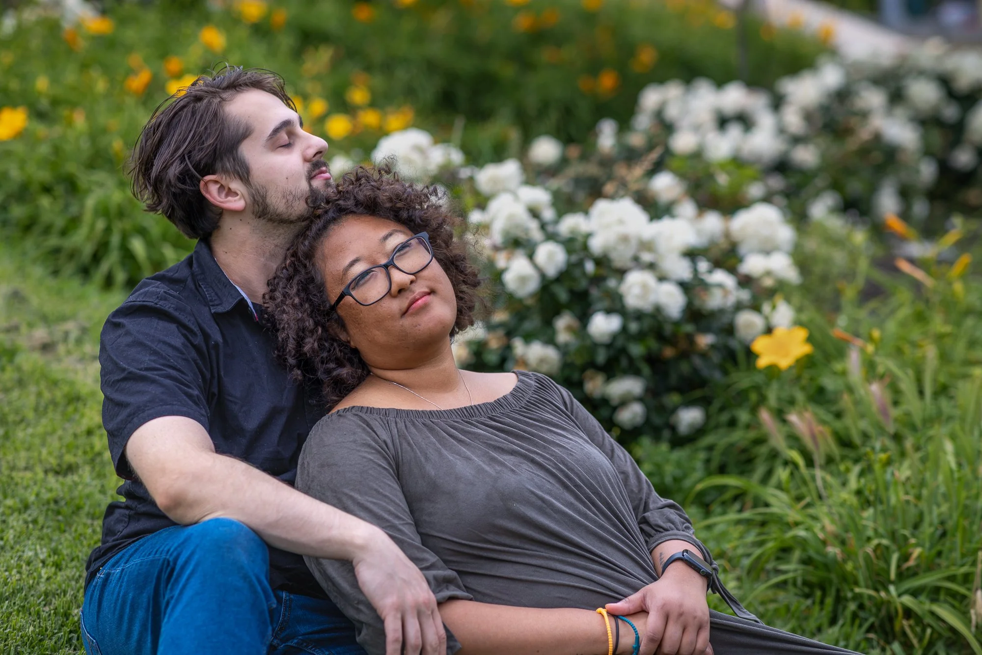 A man and woman are sitting outdoors in a garden, with the man leaning back with his eyes closed, and the woman resting her head on his chest, both appearing relaxed and content.
