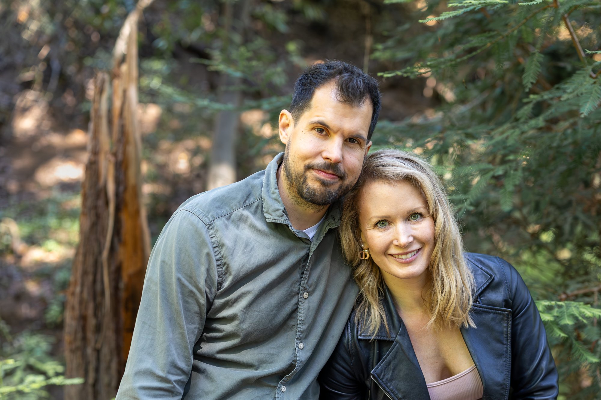 A smiling couple in a forest, with the man having dark hair and a beard, and the woman with blonde hair and green eyes, wearing a leather jacket.