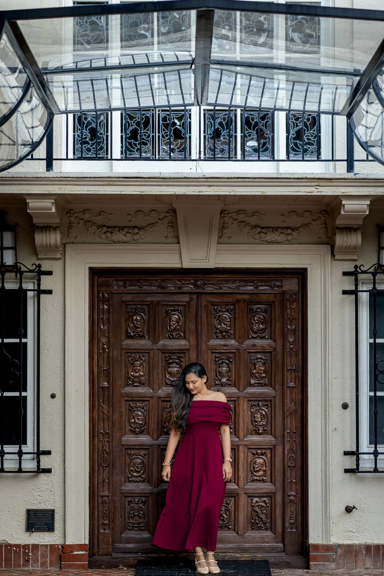 Woman in burgundy dress at historic carved wooden door of Villa Montalvo main entrance, glass canopy above, Saratoga California