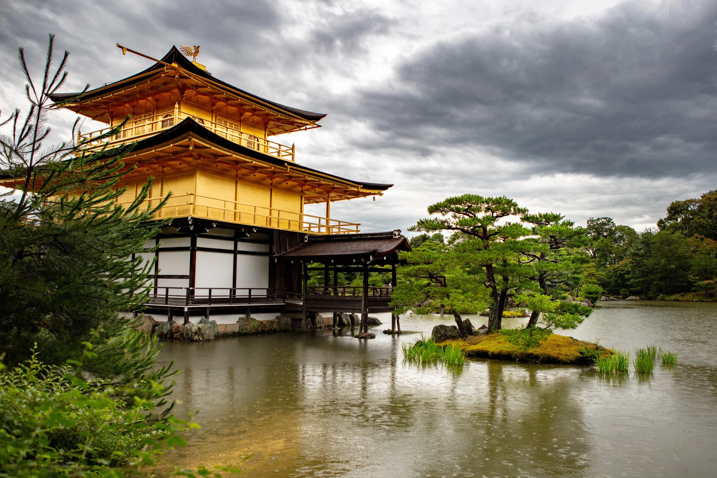 A traditional Japanese garden with a golden pavilion on a lake, surrounded by trees and a cloudy sky.