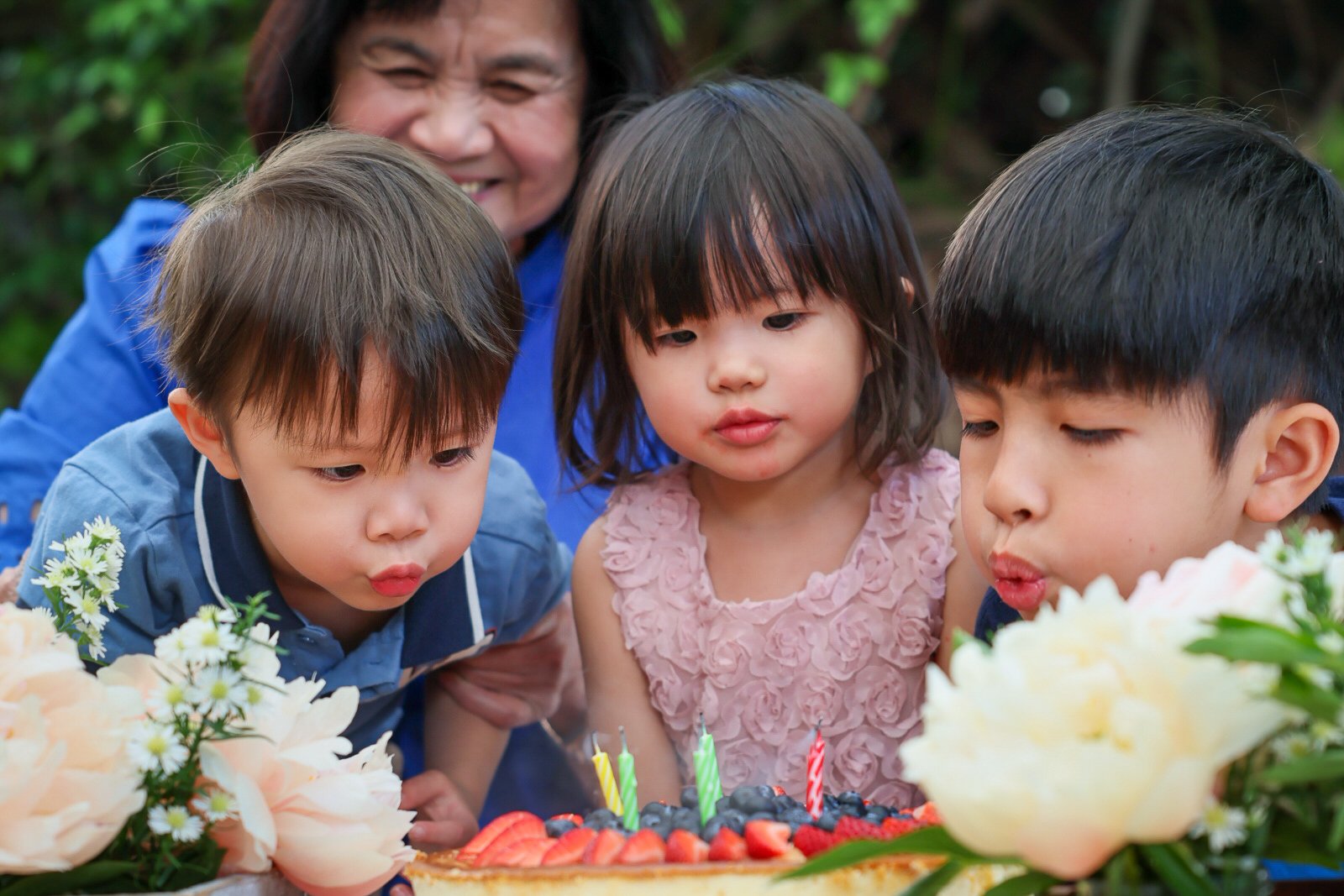 Three children and an older woman celebrating a birthday outdoors with a cake topped with berries and colorful candles, surrounded by flowers.