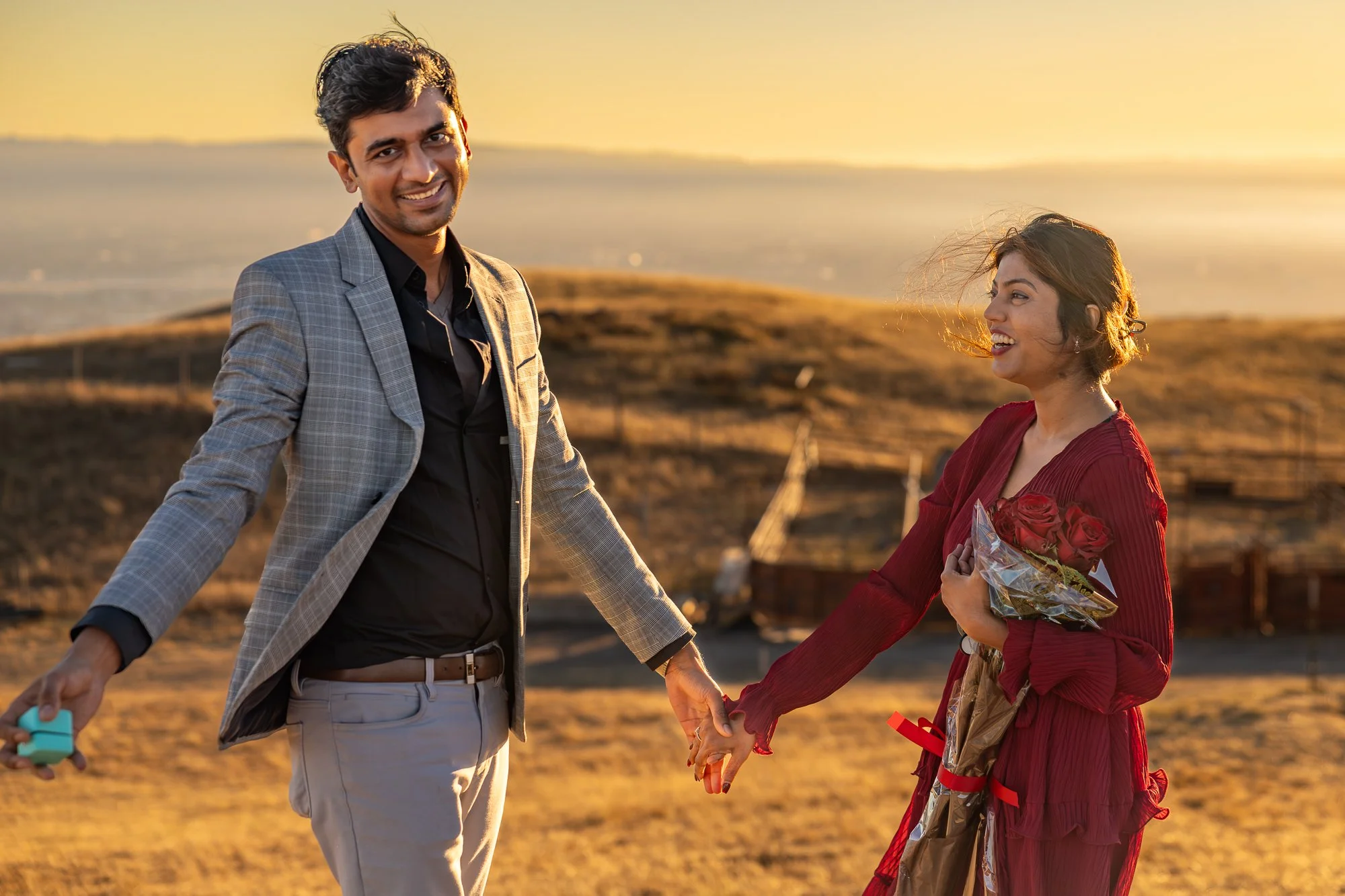 A happy couple holding hands outdoors during sunset, with the woman holding a bouquet of roses wrapped in plastic, in a field with water and hills in the background.