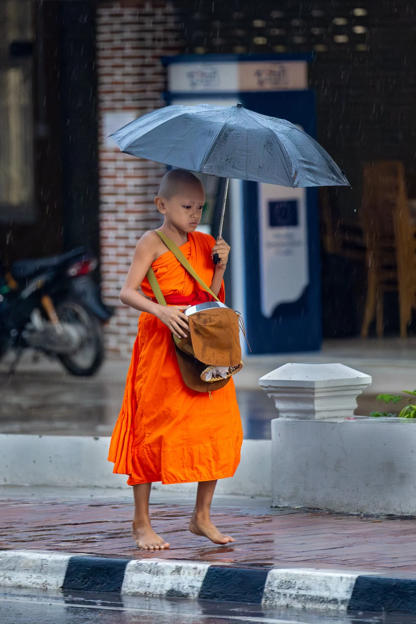 Young novice monk with grey umbrella carrying an alms bag barefoot in rain, Luang Prabang