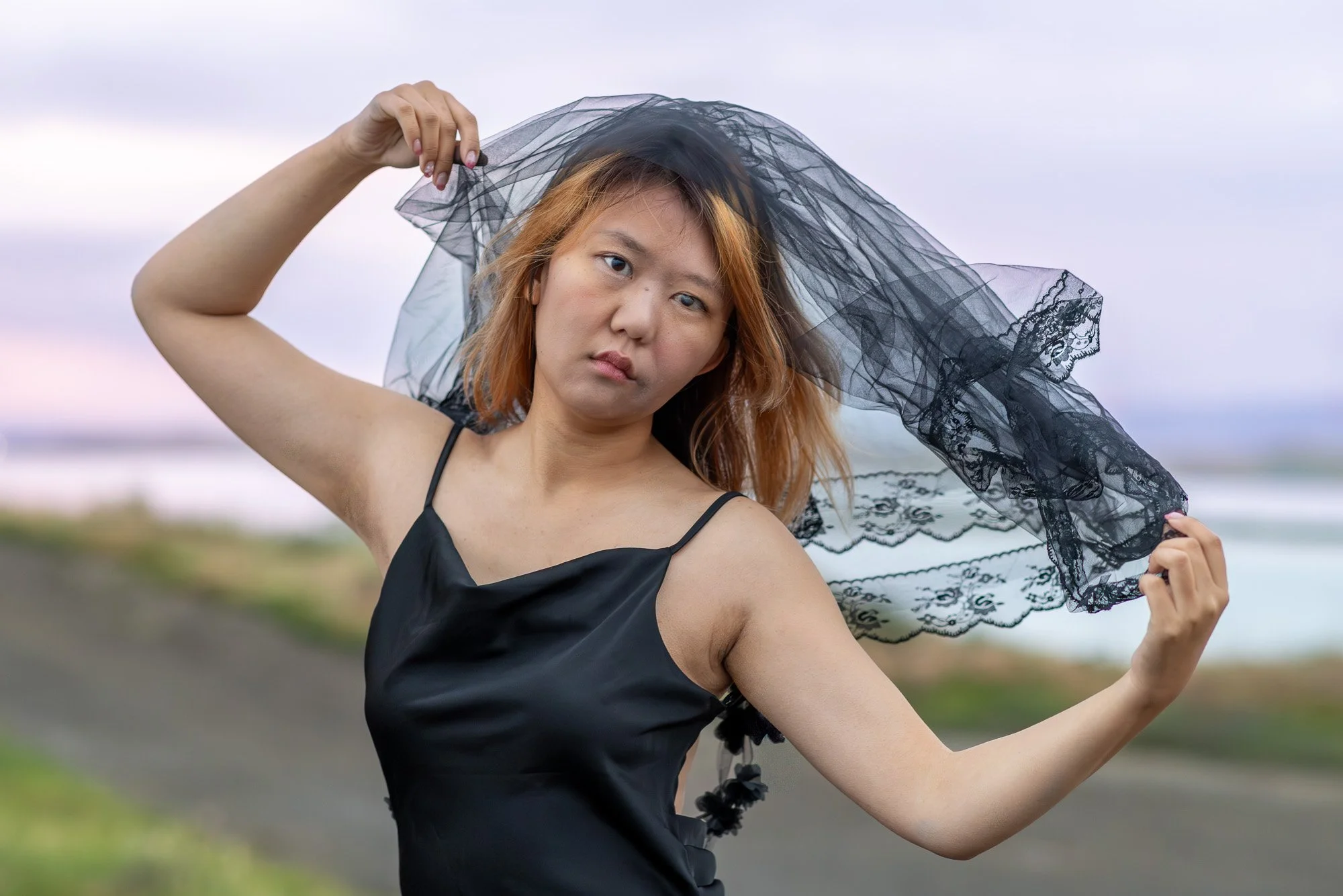 Woman in black dress holding black lace veil open wide with direct gaze, purple pink dusk sky, Dumbarton Bridge Bay Area lifestyle photography