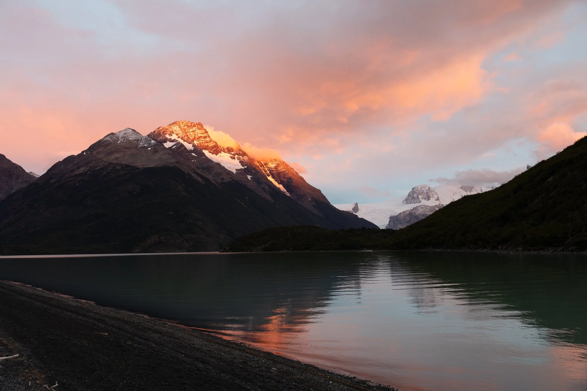 Dramatic alpenglow sunset with orange-pink light on snow-capped mountains reflected in glacial lake at Refugio Seron, Torres del Paine