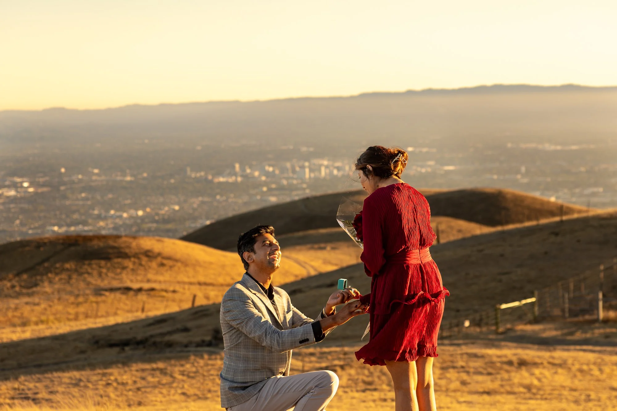 A man on one knee proposing marriage to a woman in a red dress during sunset on a hilly landscape with city in the background.