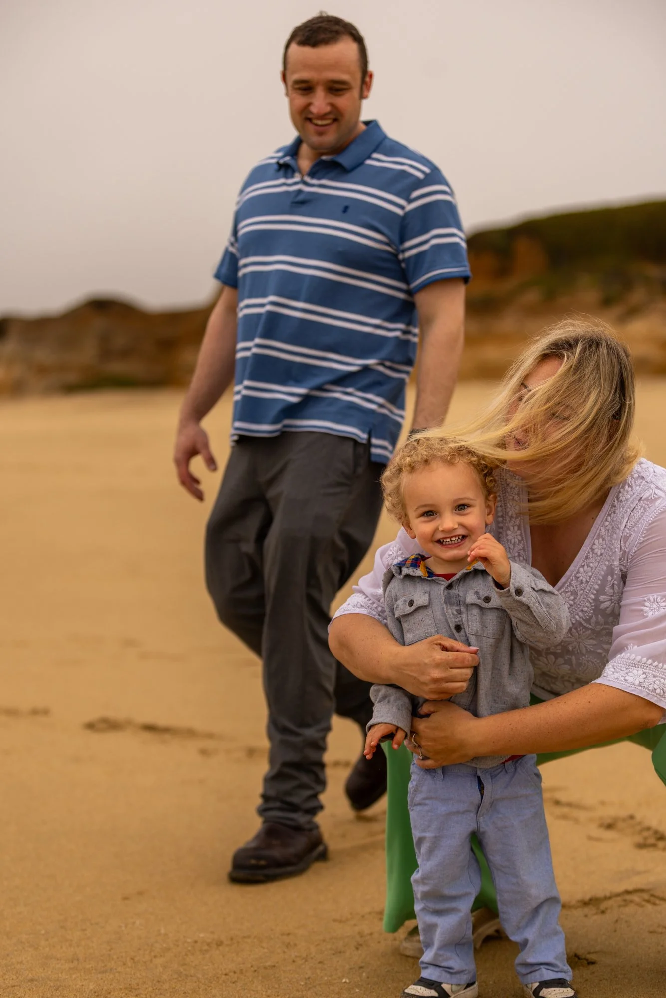 A happy woman holds a young boy with curly hair in her arms on a sandy beach, smiling at the camera. An adult man in a blue striped polo shirt and khaki pants stands behind them, smiling and looking down. There are sand dunes and a cloudy sky in the 