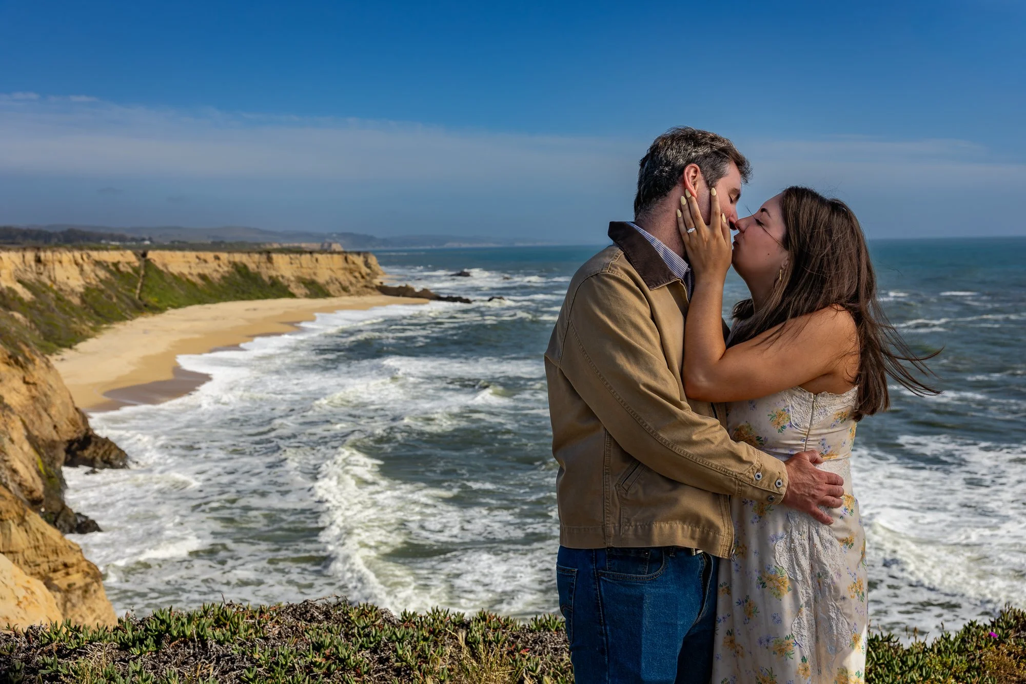 A couple kissing by the ocean on a sunny day, with cliffs and waves in the background.