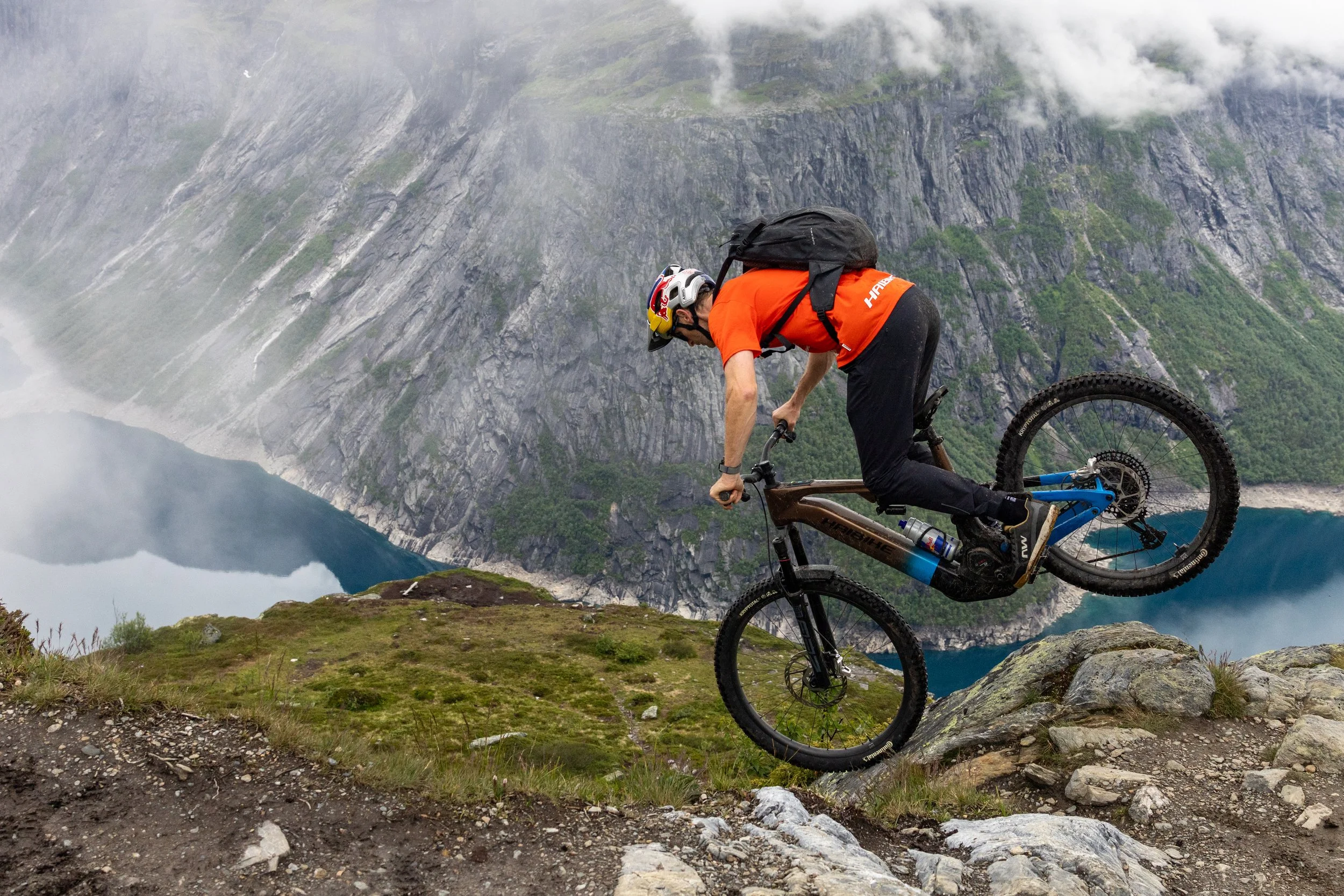 Mountain biker in orange shirt riding downhill on rocky trail with mountains and a lake in the background.