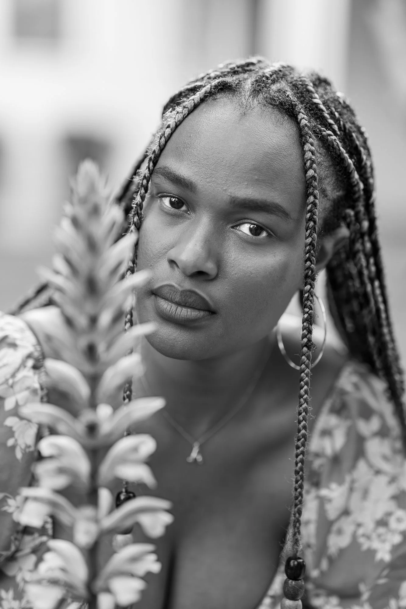 Black and white close-up portrait of a woman with braided hair, gazing at the camera amidst plants in the foreground.