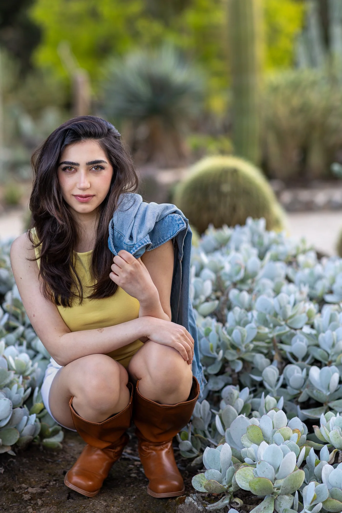 Woman in yellow top with denim jacket off shoulder crouching among silver succulent plants at Stanford Cactus Garden, lifestyle portrait