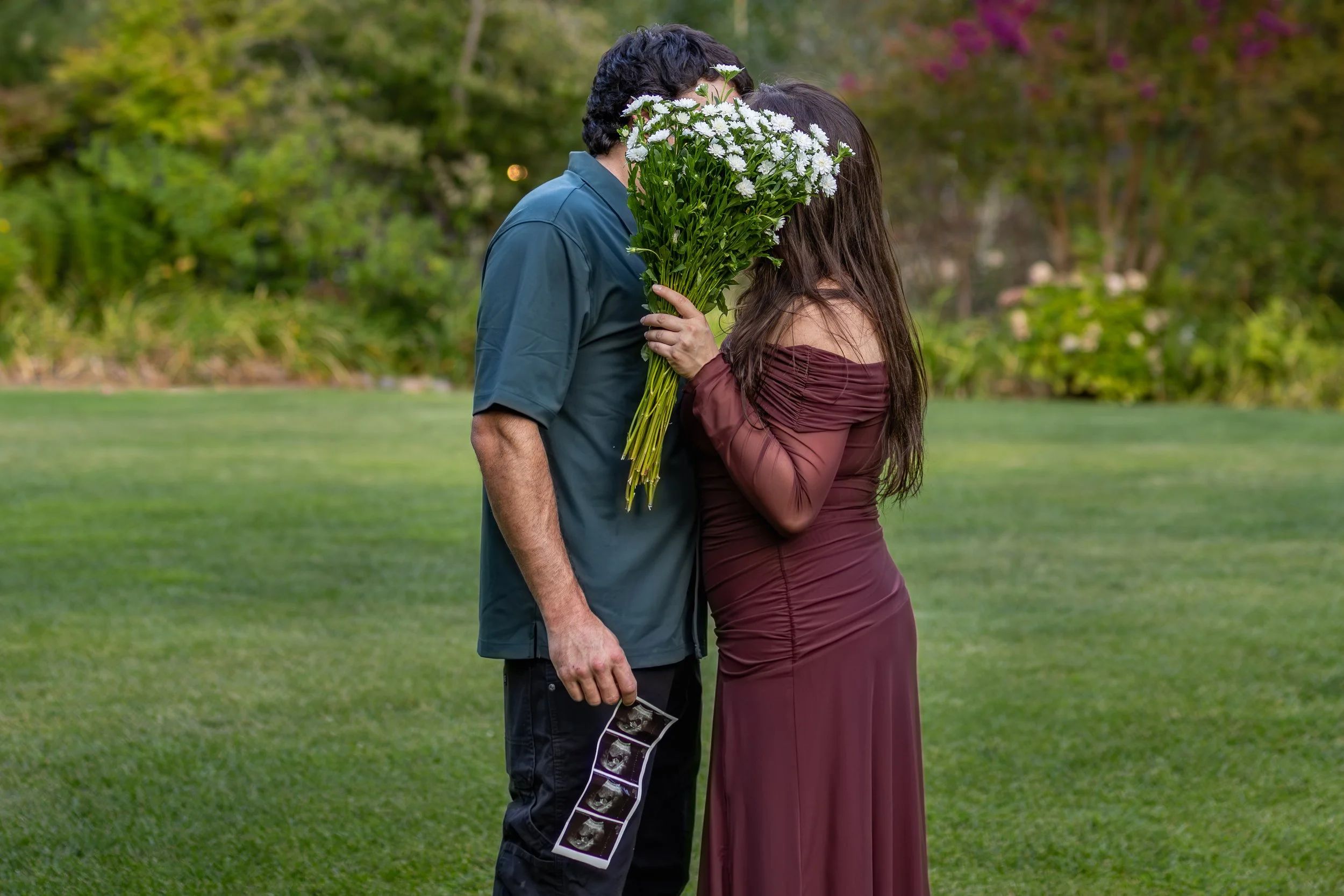 A woman in a maroon dress and a man in a blue shirt kissing outdoors, holding a large bouquet of white flowers.