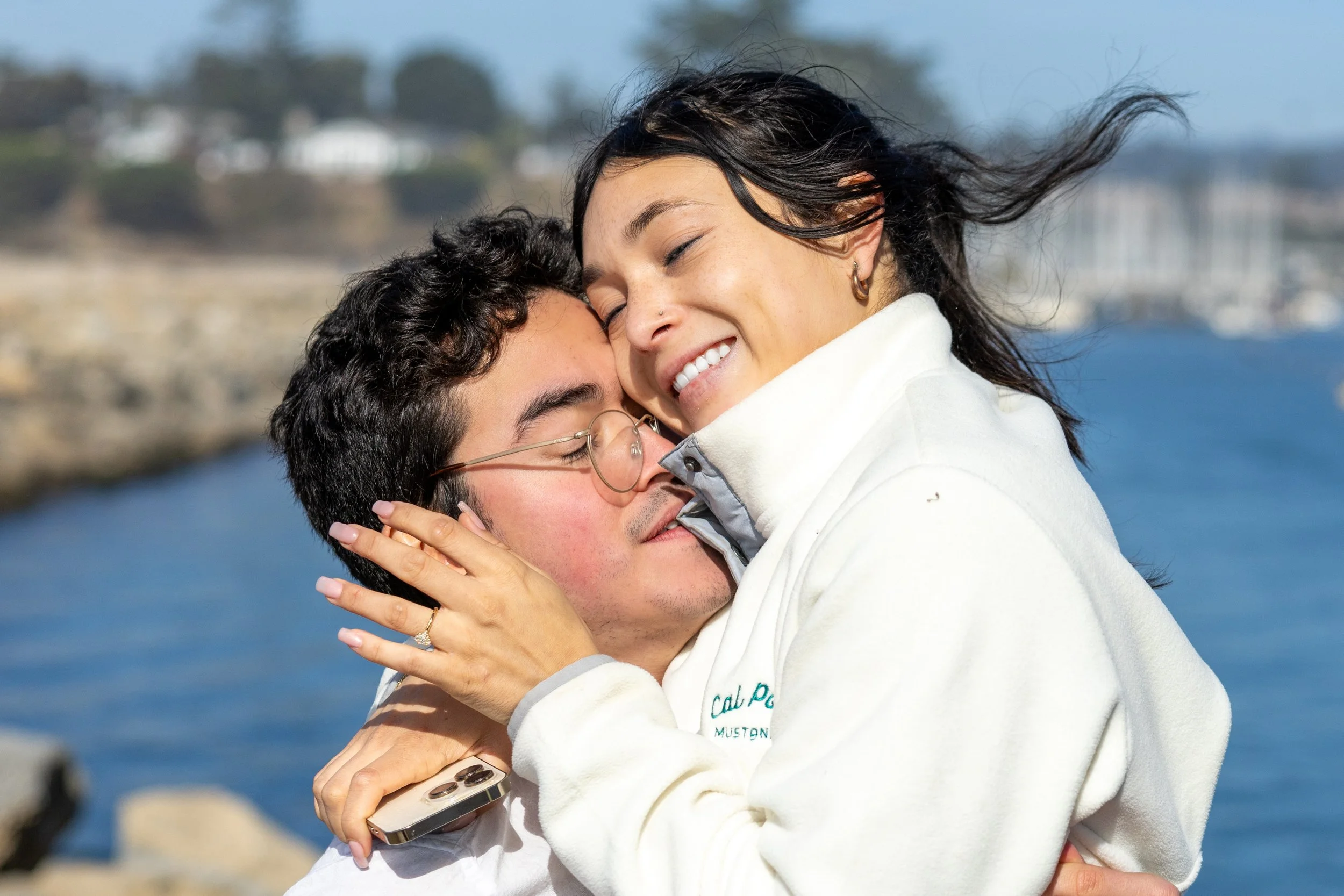A young couple embracing outdoors near a body of water, smiling with eyes closed.