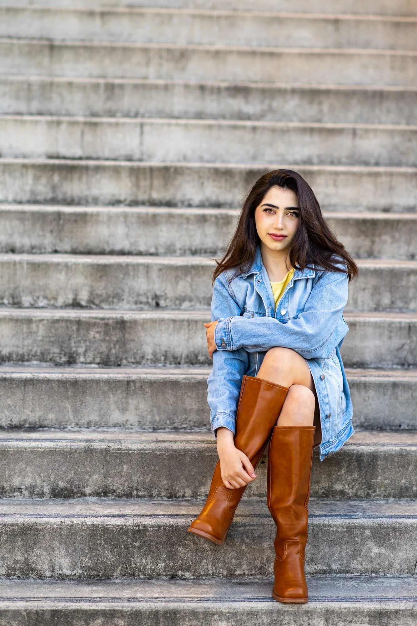 A young woman with long dark hair and light skin sitting on outdoor concrete steps, wearing a yellow top, denim jacket, shorts, and knee-high brown leather boots, holding a matching brown leather handbag.