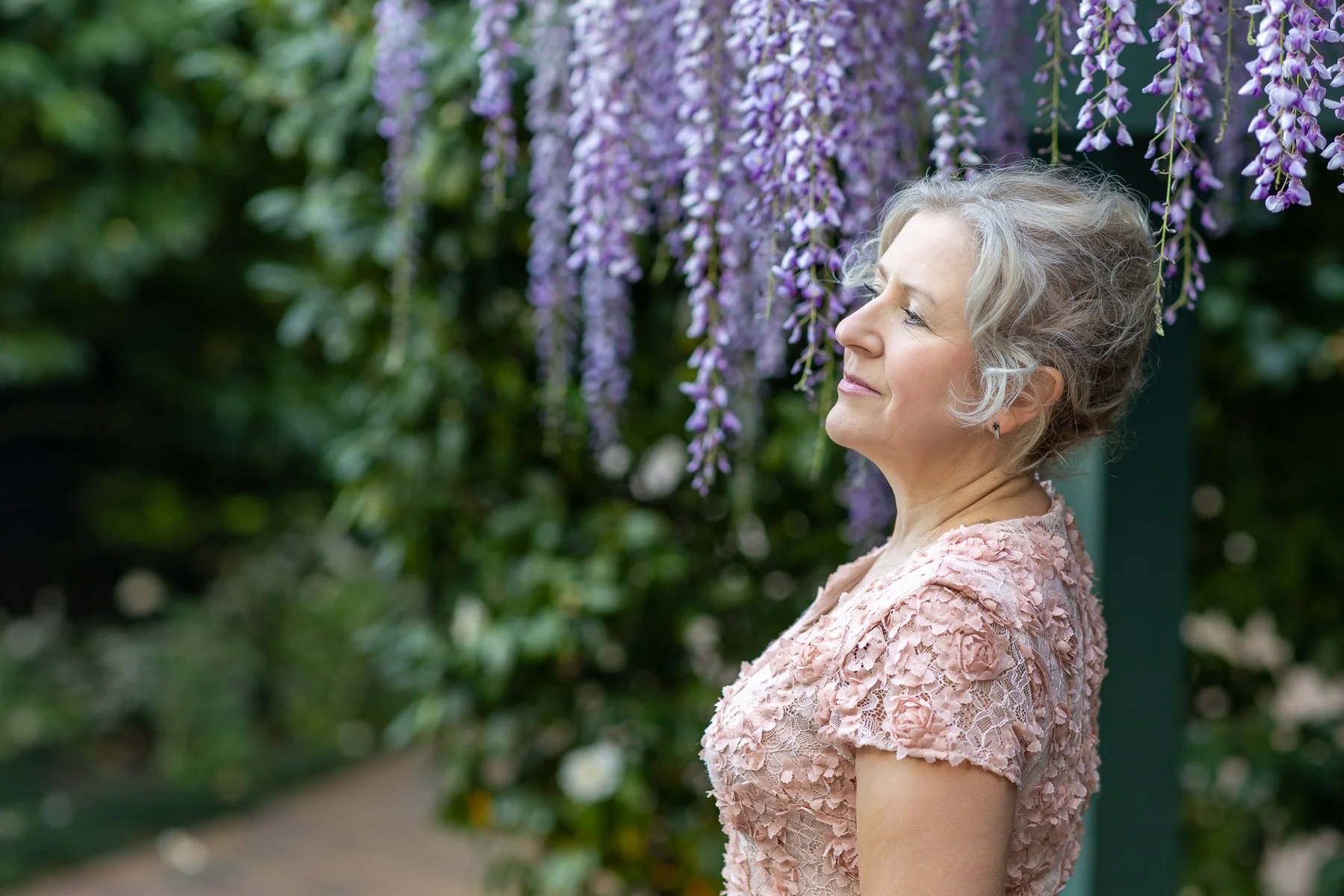 Woman in pink lace dress with eyes closed beneath purple wisteria cascades at Gamble Garden, Palo Alto, natural light portrait