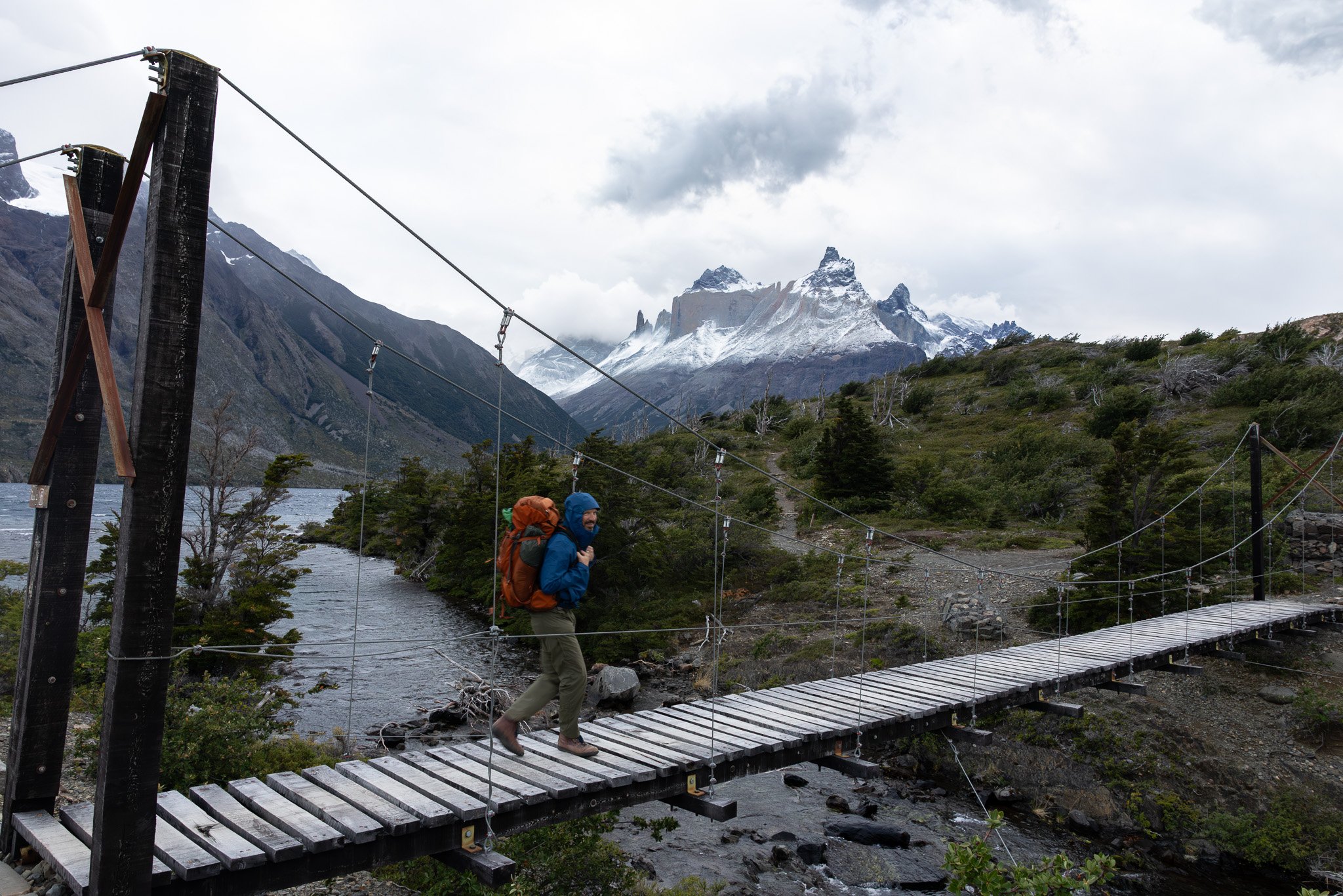 Hiker with orange backpack walking on suspension bridge away down trail directly toward snow-covered Los Cuernos massif, Torres del Paine