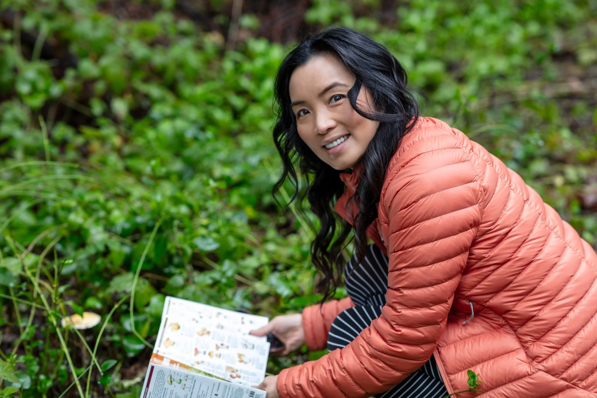 Woman in orange jacket crouching with mushroom field guide in lush green Bay Area forest, Santa Cruz Mountains winter foraging