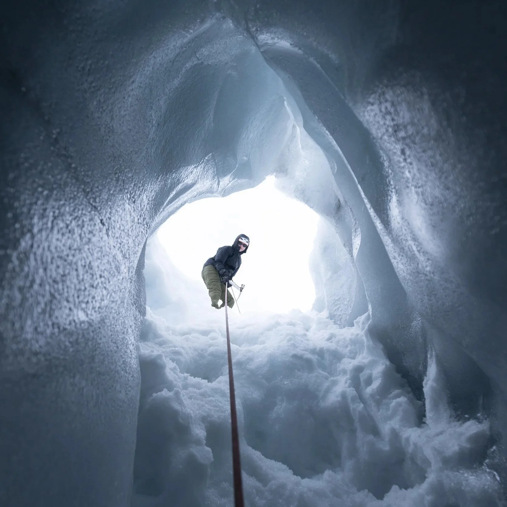 An ice climber wearing black gear, including a helmet and sunglasses, ascending inside a frozen ice cave using climbing tools and a rope.