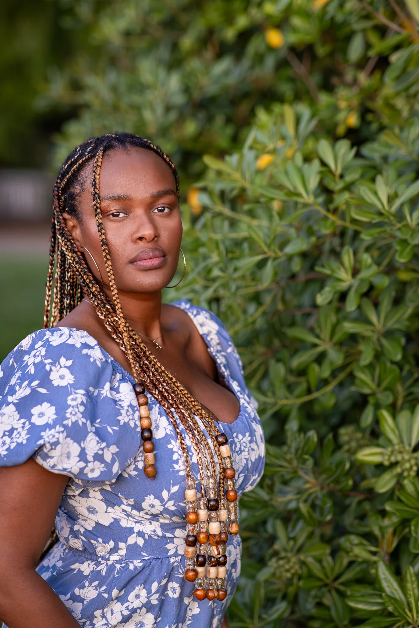 A woman with braided hair wearing hoop earrings and layered beaded necklaces standing outdoors in front of green foliage, wearing a blue floral dress.