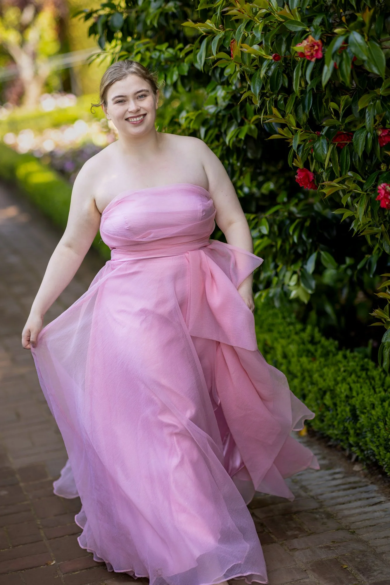 A young woman in a pink strapless dress walking outdoors along a brick path, surrounded by green bushes and plants, with sunlight filtering through trees.