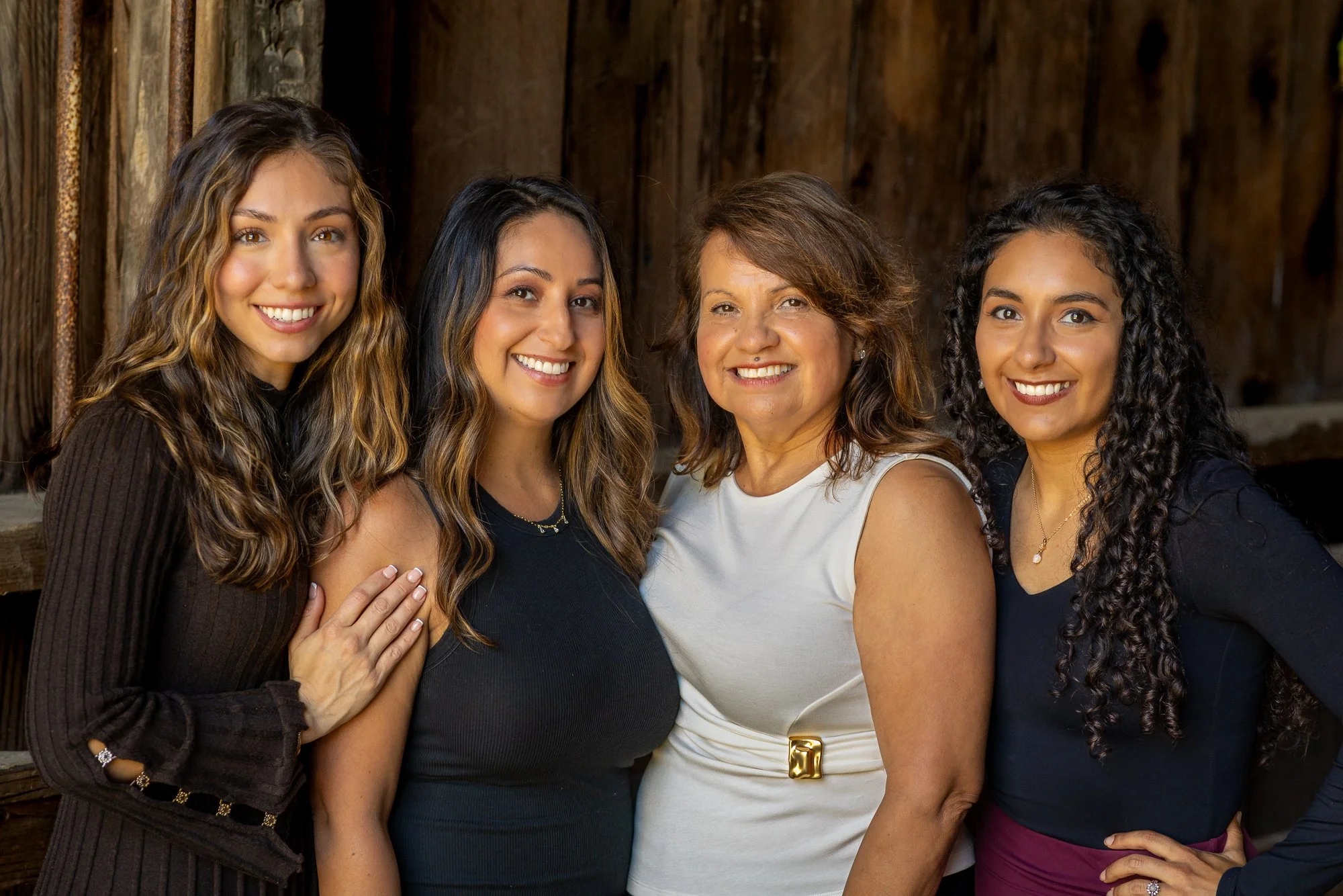 Four women with different hair colors and styles smiling together in front of a wooden background, posing closely with arms around each other.