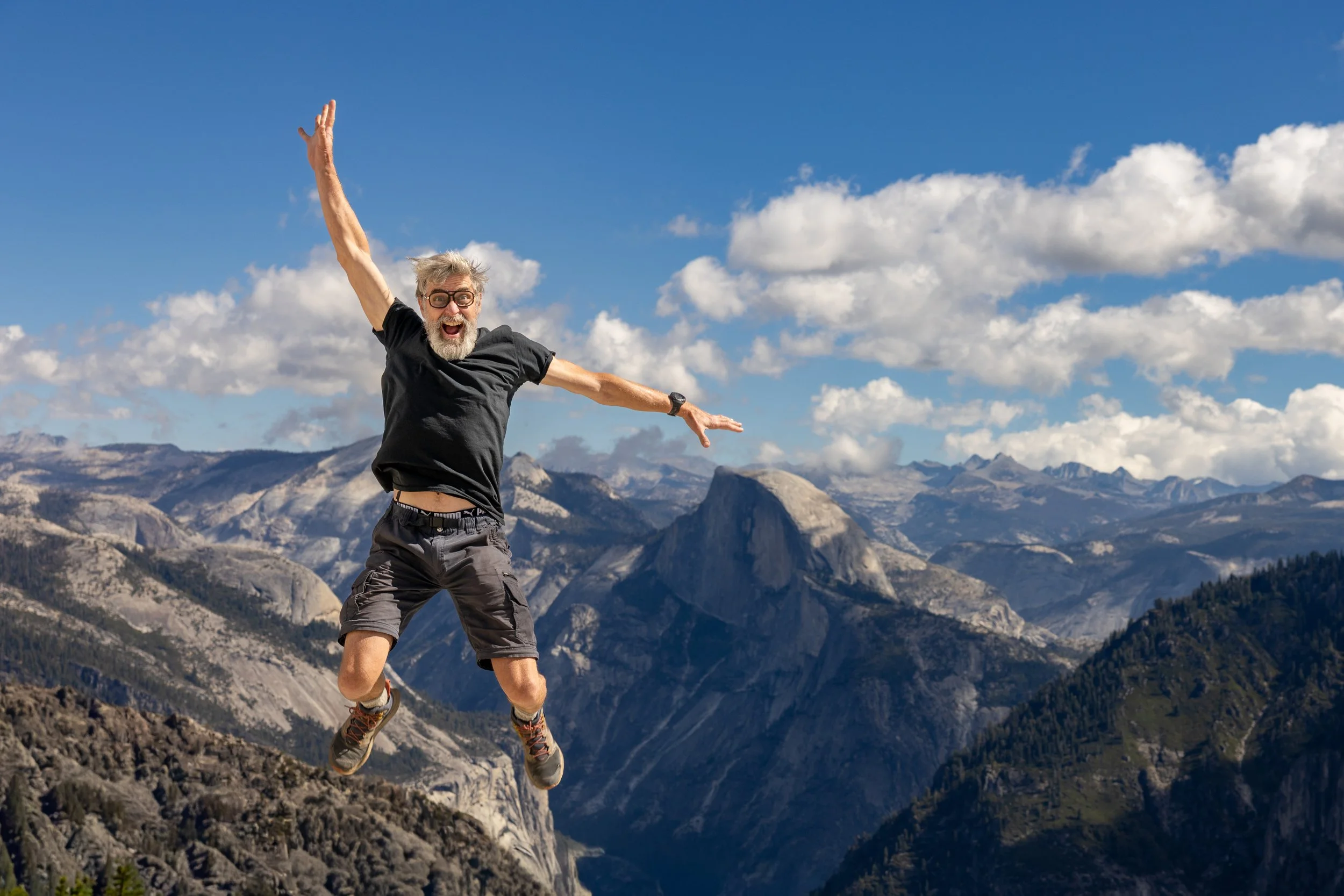 A man with gray hair, beard, glasses, wearing a black t-shirt and shorts, is jumping in the air with a happy expression against a backdrop of mountainous terrain and a partly cloudy blue sky.