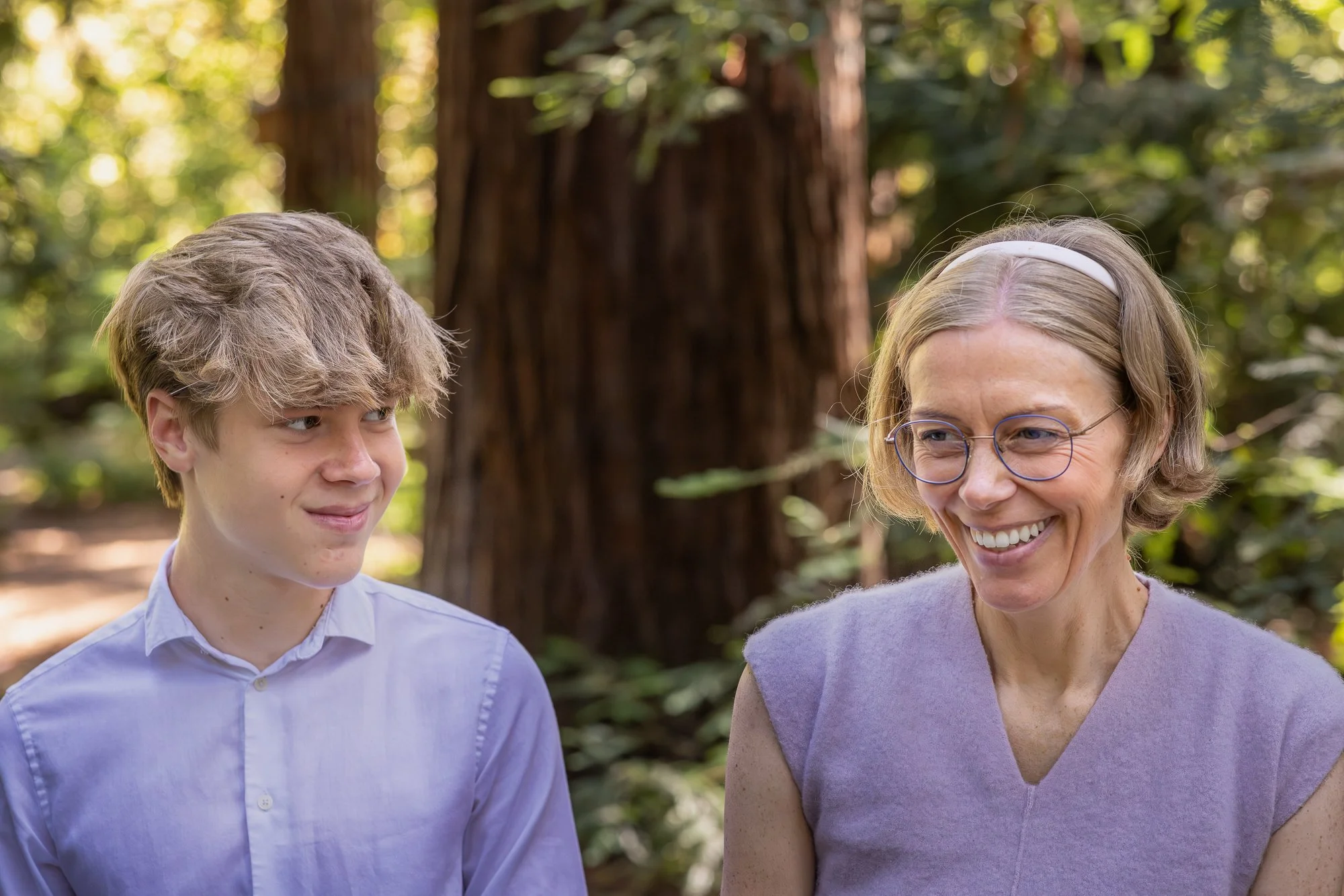 A young man with light brown, tousled hair and light skin, wearing a white button-up shirt, standing outdoors in a forested area. Beside him, a middle-aged woman with shoulder-length blonde hair, glasses, and light skin, wearing a lavender short-slee