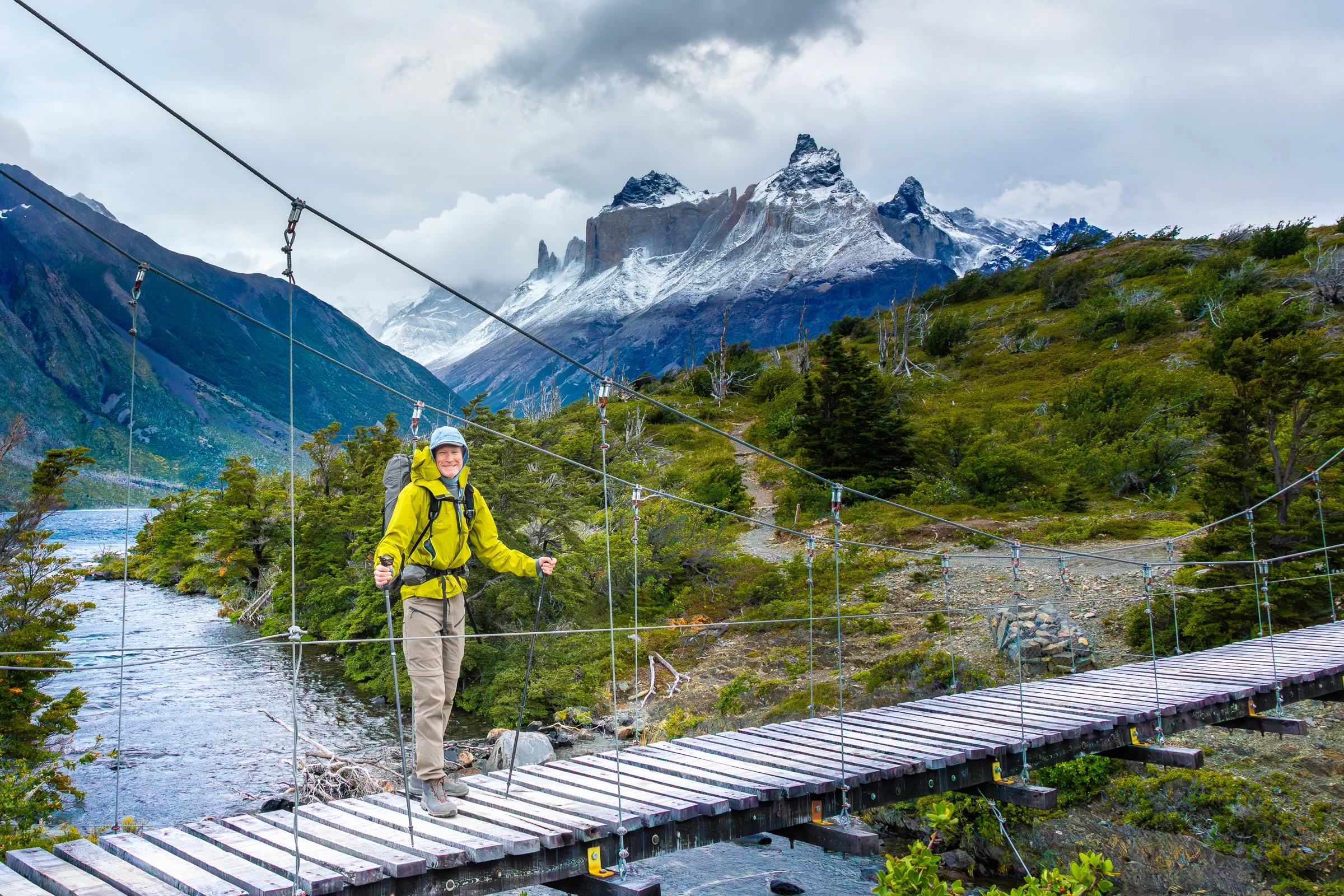 A person wearing a yellow jacket, beige pants, and a helmet, hiking across a suspension bridge over a river in a mountainous area with snow-capped peaks and greenery.