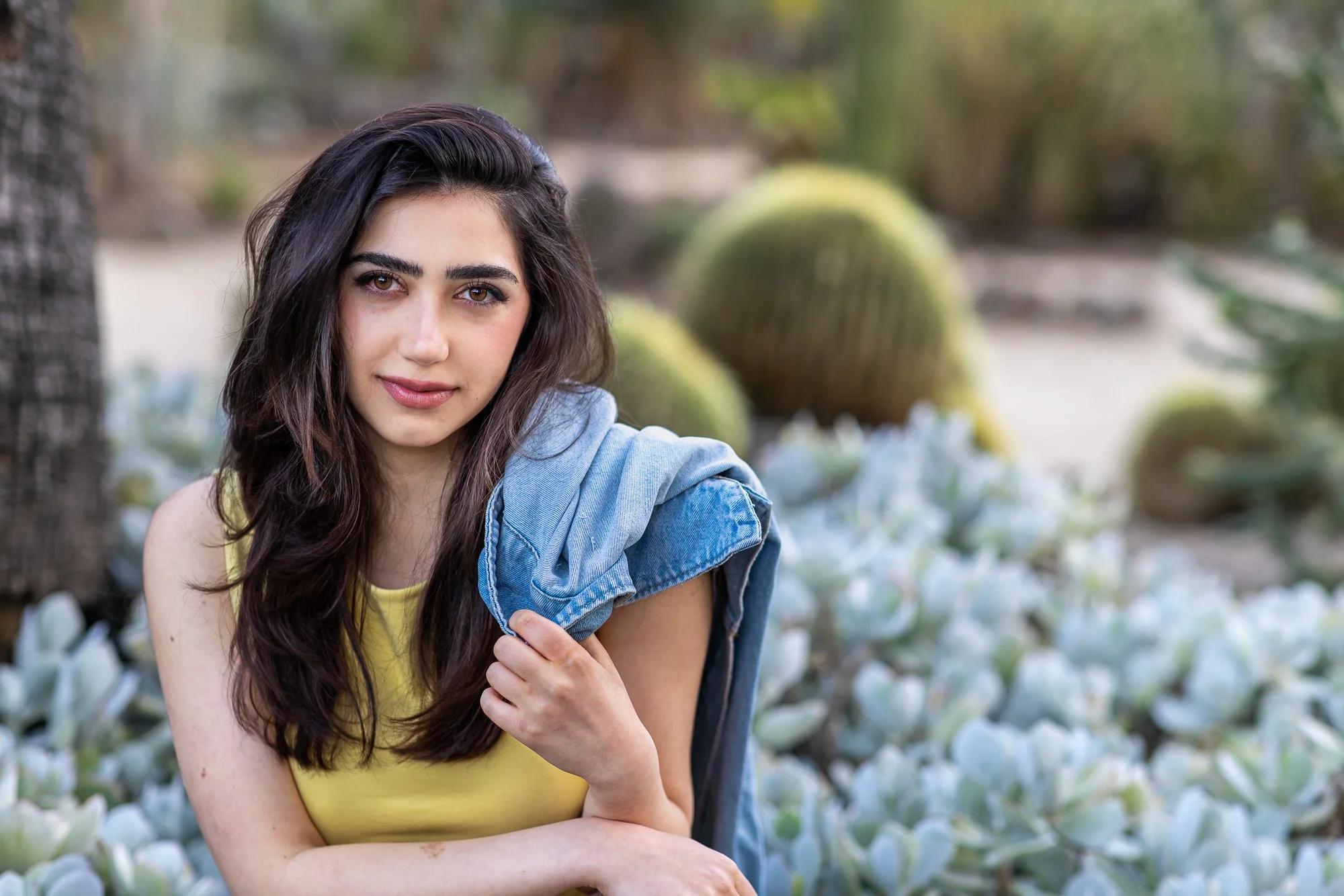 A young woman with long dark hair and hazel eyes, wearing a yellow top and a denim jacket draped over her shoulder, sitting outdoors amidst white flowering plants and large round green cacti, with a blurred natural background.