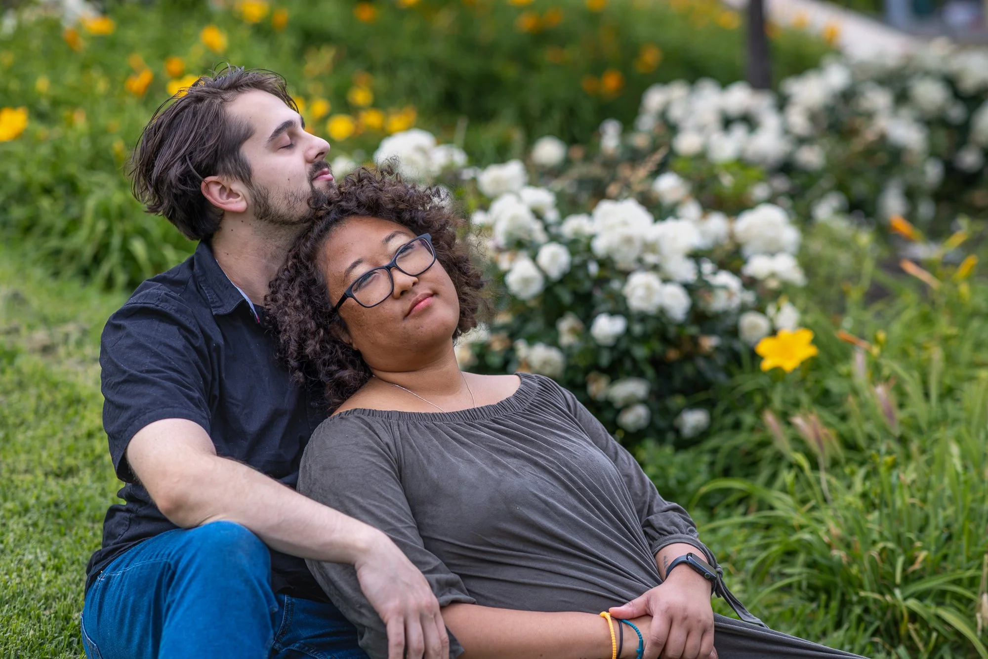 A young man with dark hair and beard, eyes closed, sitting behind a woman with curly hair and glasses, resting against him in a garden with green grass and blooming white and yellow flowers.