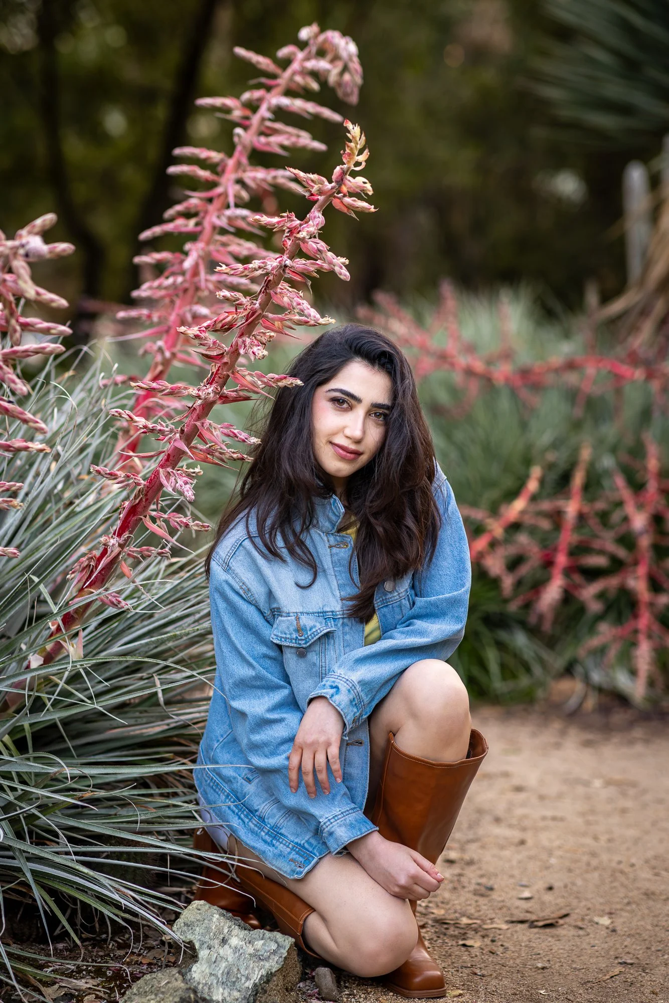A young woman with long dark hair, wearing a denim jacket and brown knee-high boots, kneeling on the ground amidst large pink flowering plants and greenery in an outdoor setting.