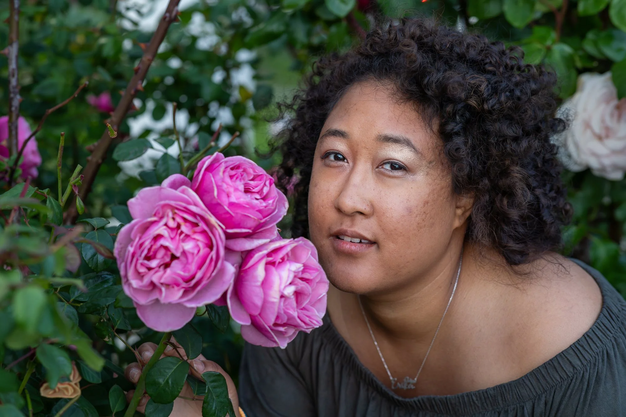 A woman with curly dark hair and a necklace with the name 'Enrique' is standing close to pink and white roses in a garden.
