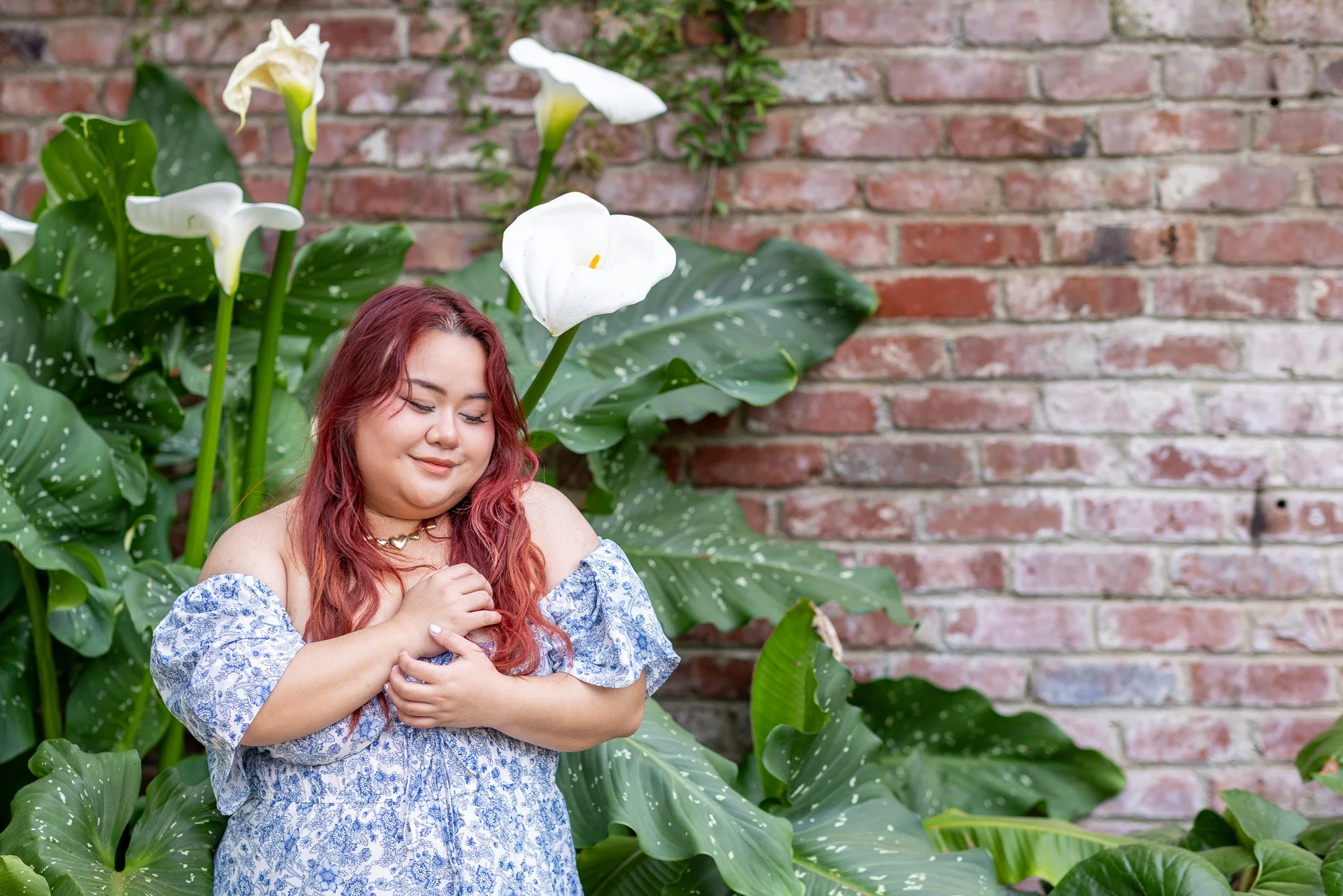 Woman in blue floral dress with eyes closed and hands at chest beside white calla lilies against brick wall, meditative portrait Elizabeth Gamble Garden Palo Alto