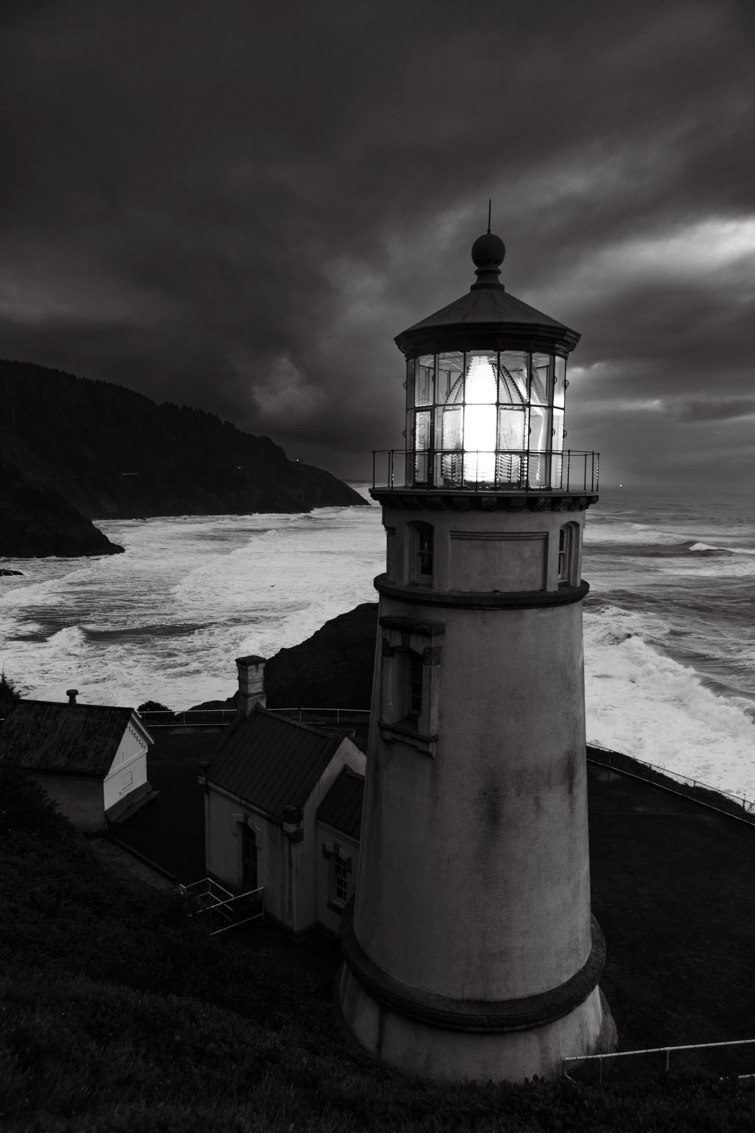 Black and white photo of a lighthouse by the ocean with stormy sky, waves crashing on the shore, and cliffs in the background.