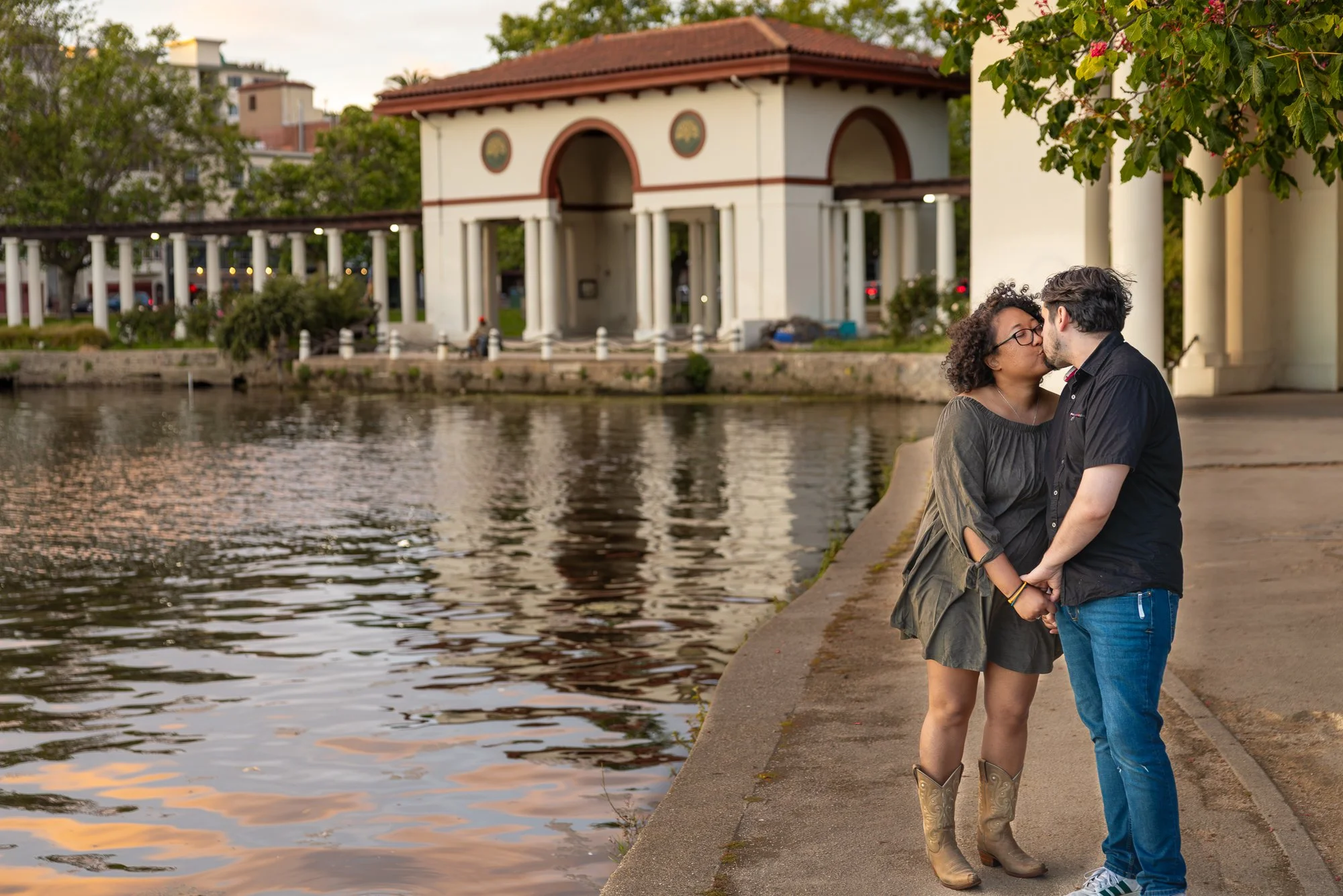 A couple sharing a kiss by the riverwalk with a white pavilion in the background, trees, and buildings, during sunset.