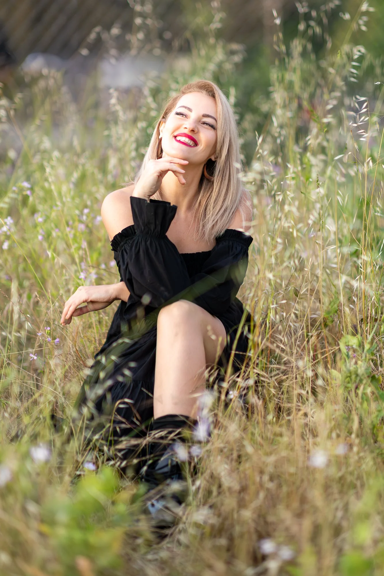 A joyful woman with blonde hair and red lipstick smiling and posing in a field of tall grass and wildflowers.