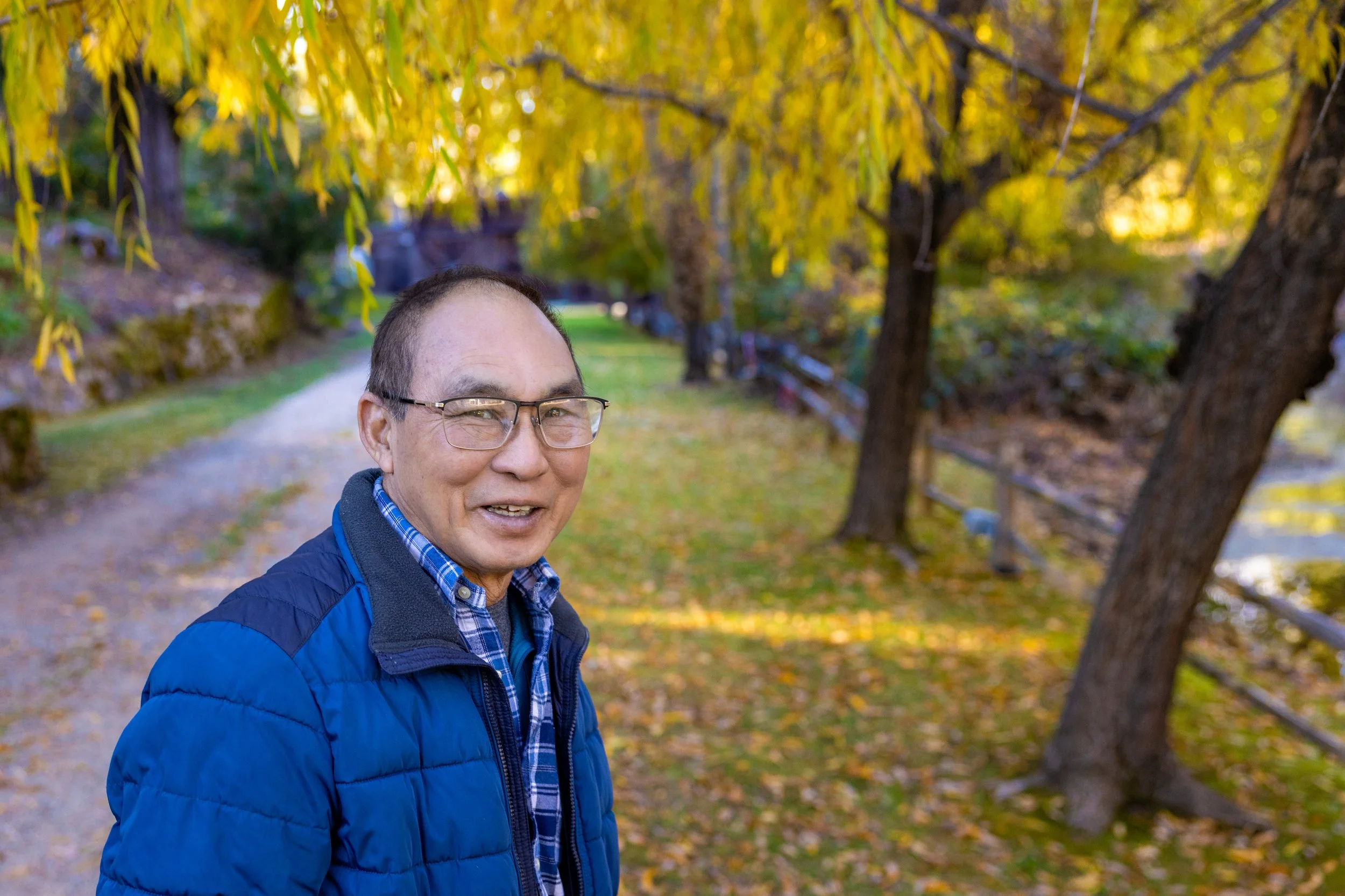 An older man with glasses and a blue jacket smiling outdoors during autumn, standing on a path surrounded by yellow and green fall foliage.