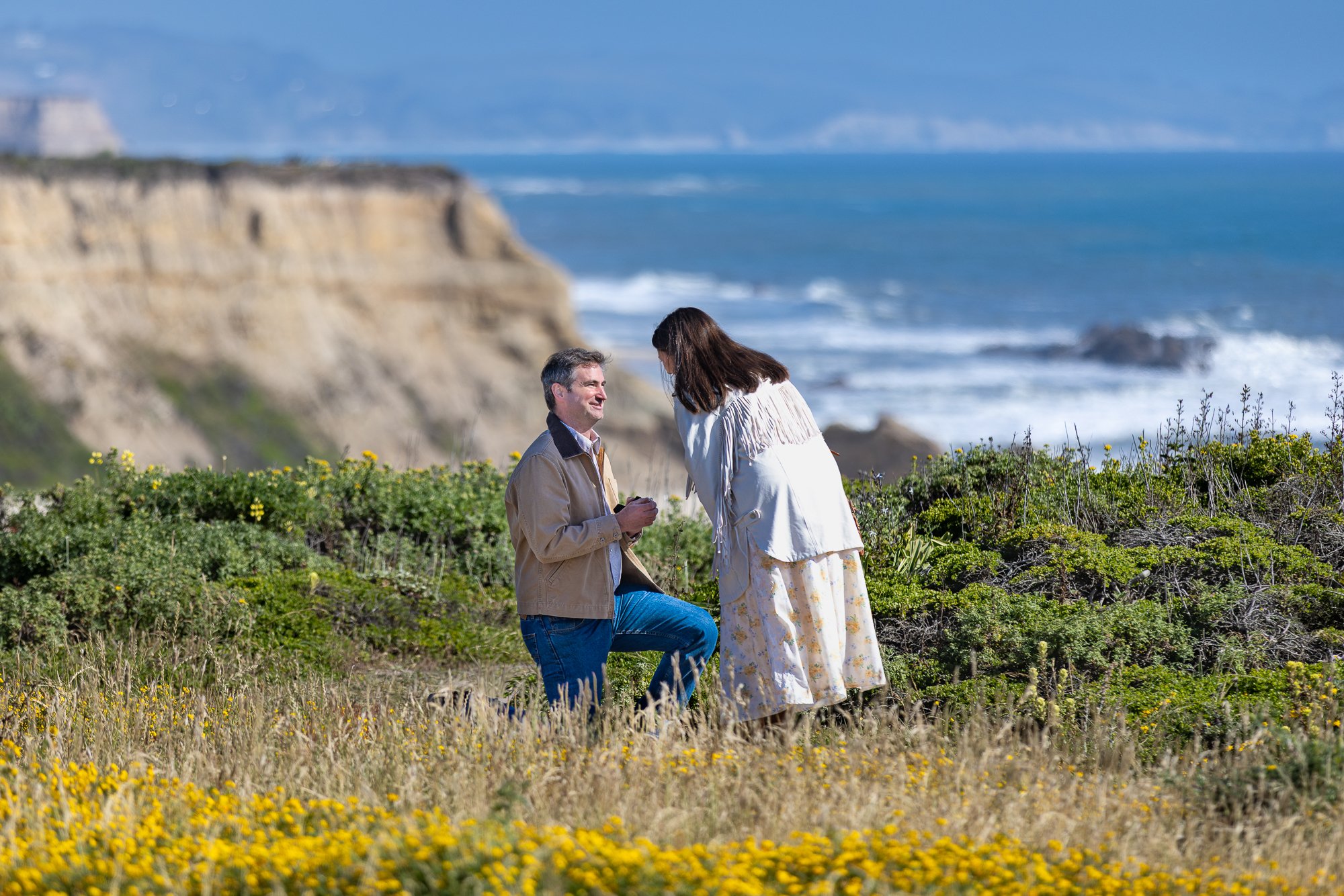 A man kneeling on one knee and proposing marriage to a woman in a field of yellow flowers near the coast, with cliffs and ocean in the background.