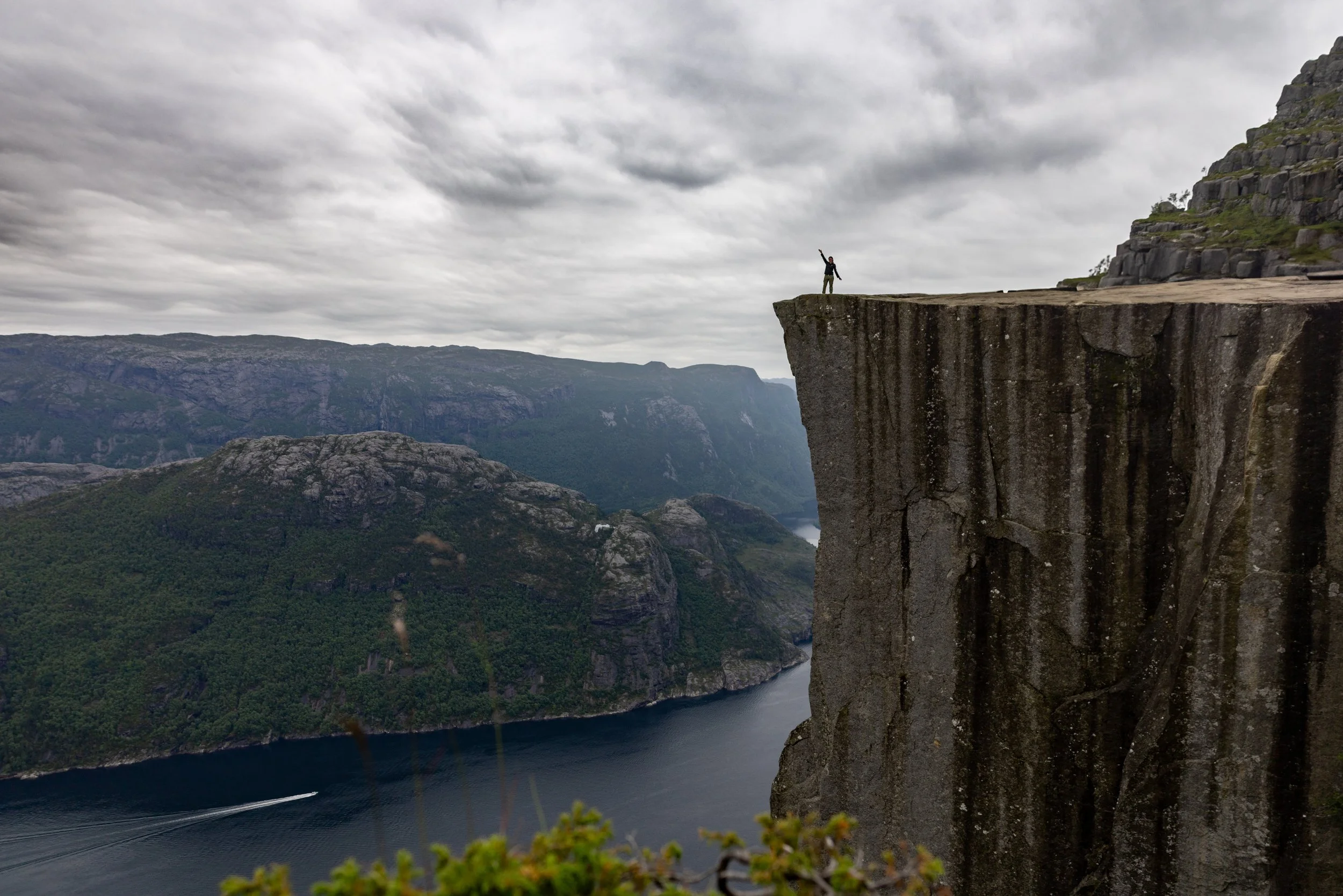 A person standing at the edge of a large cliff with a raised arm in a scenic mountain landscape under cloudy sky.
