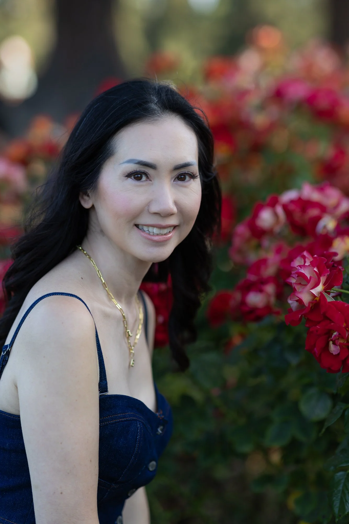 A woman with dark hair and light skin smiling, wearing a dark blue top with thin straps, a gold necklace, and makeup, standing outdoors in front of pink and red flowering plants.