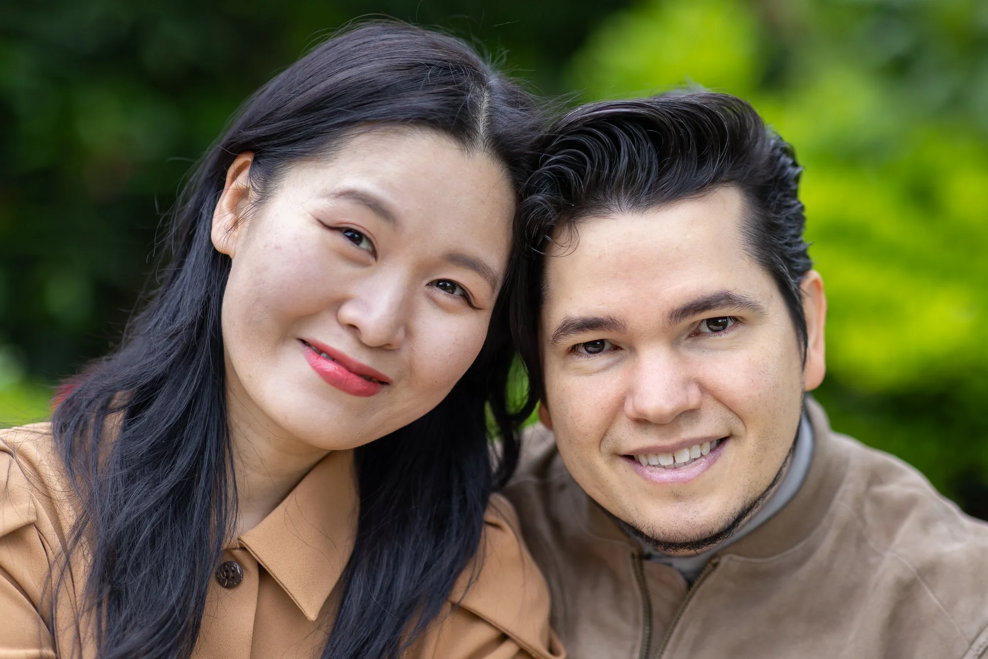 A woman and a man smiling outdoors with green foliage in the background, close up of their faces.
