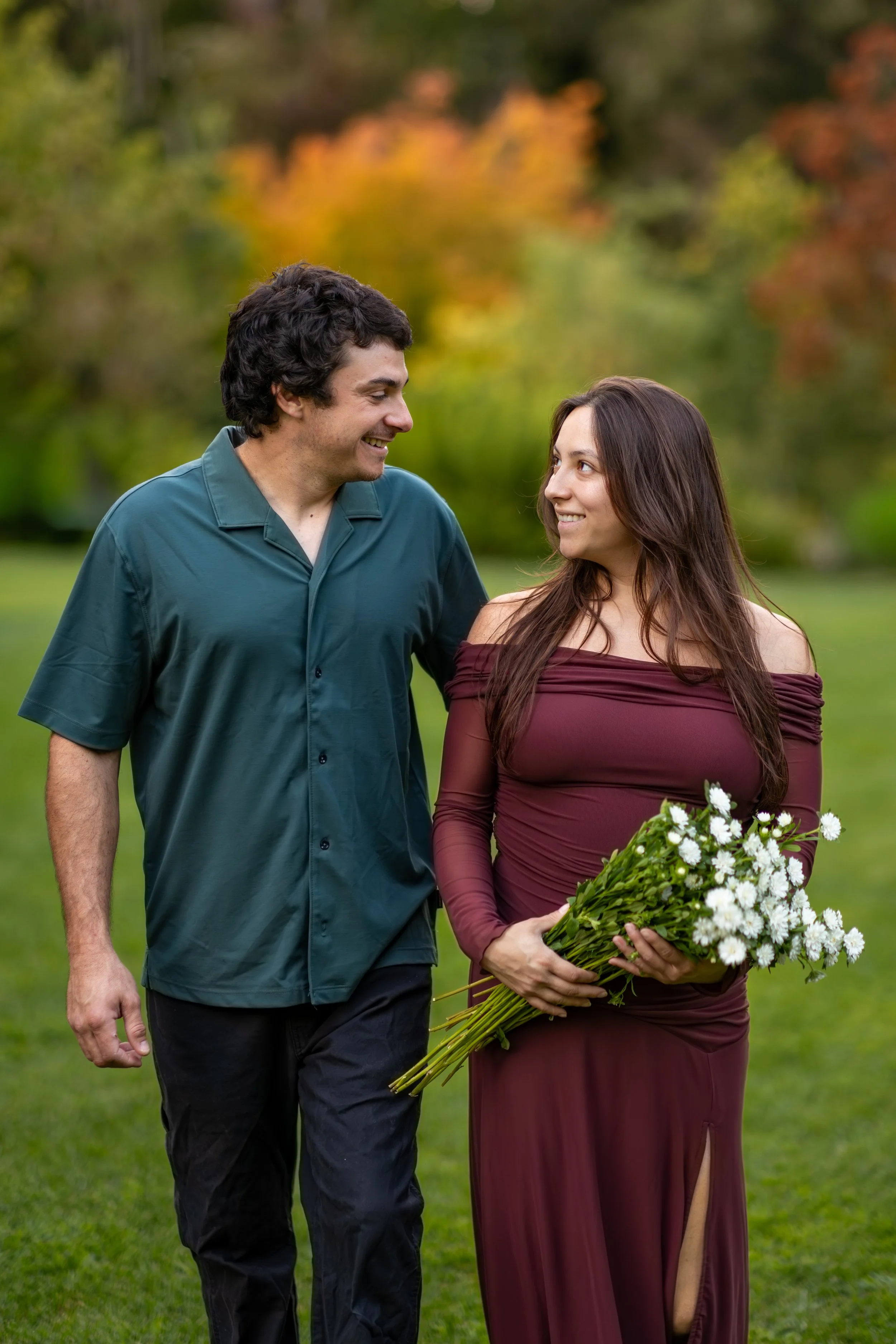A man and woman standing outdoors on a grassy area with autumn-colored trees in the background. The man has dark, curly hair and is smiling at the woman. The woman has long, brown hair and is holding a bouquet of white flowers, wearing a maroon off-t