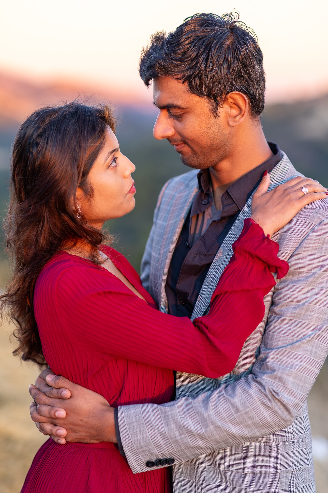 A man and woman are embracing outdoors during sunset, looking into each other's eyes. The woman is wearing a red dress with ruffled sleeves, and the man is dressed in a light gray plaid blazer with a dark shirt underneath. The woman has her hands on 