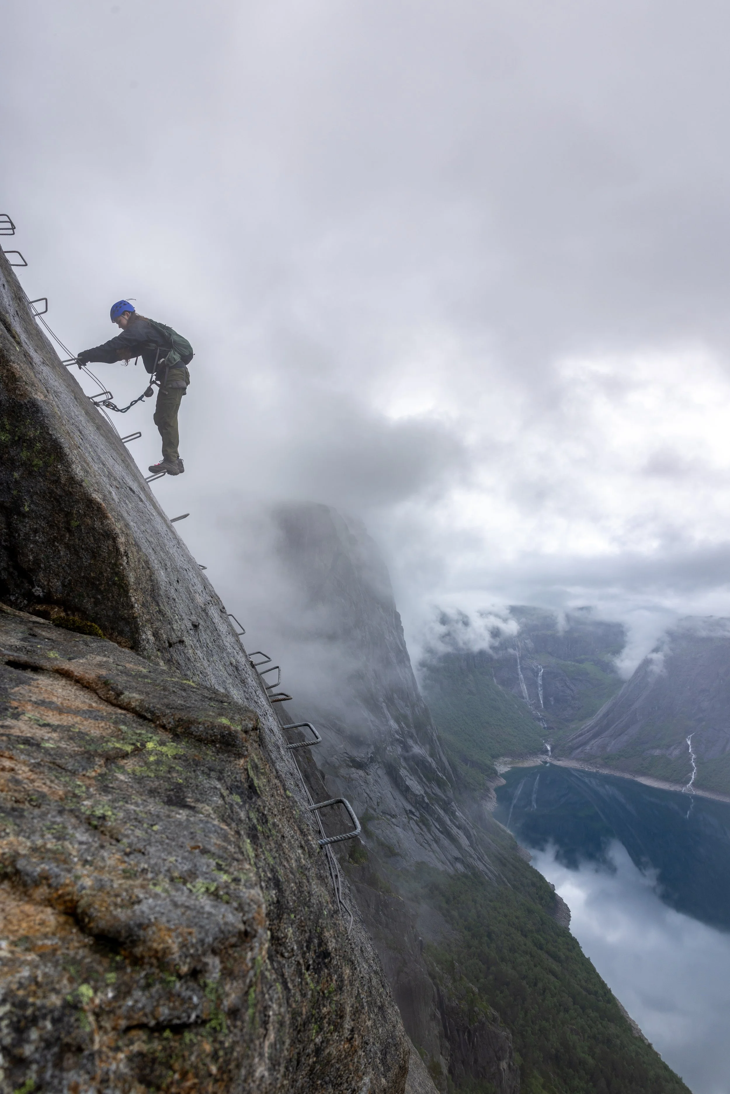 A person wearing a blue helmet and harness climbing a steep rock face with safety cables in a mountainous area with fog and clouds, overlooking a fjord.